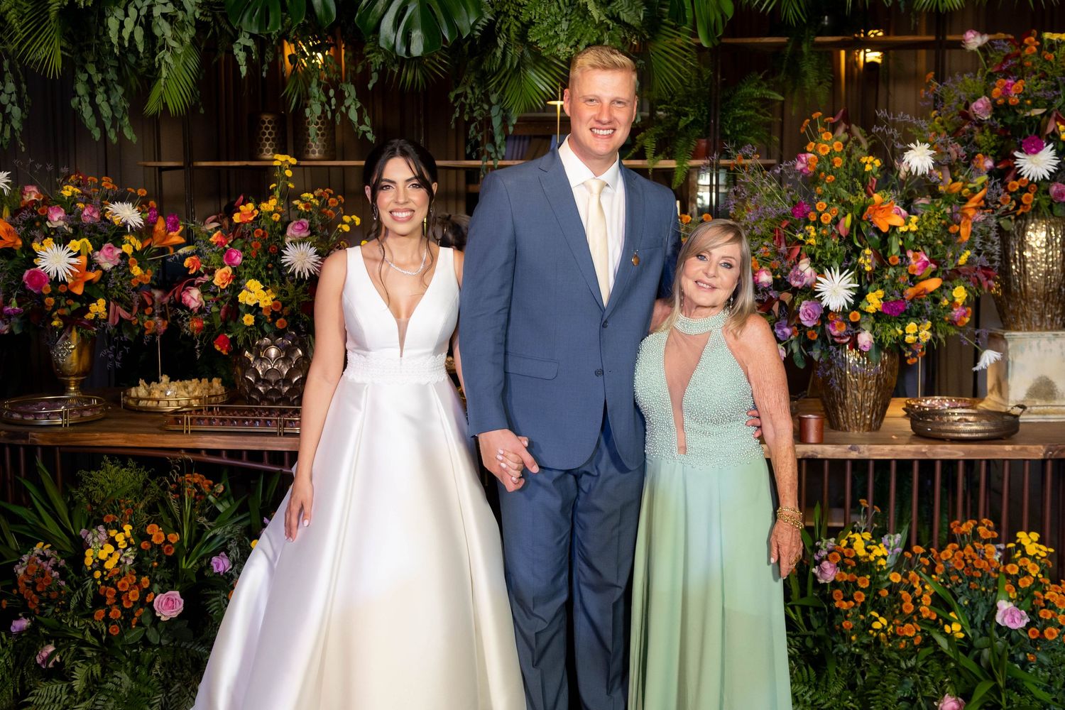 Bride in white gown, groom in blue suit, and woman in mint dress pose before a floral backdrop at a wedding.