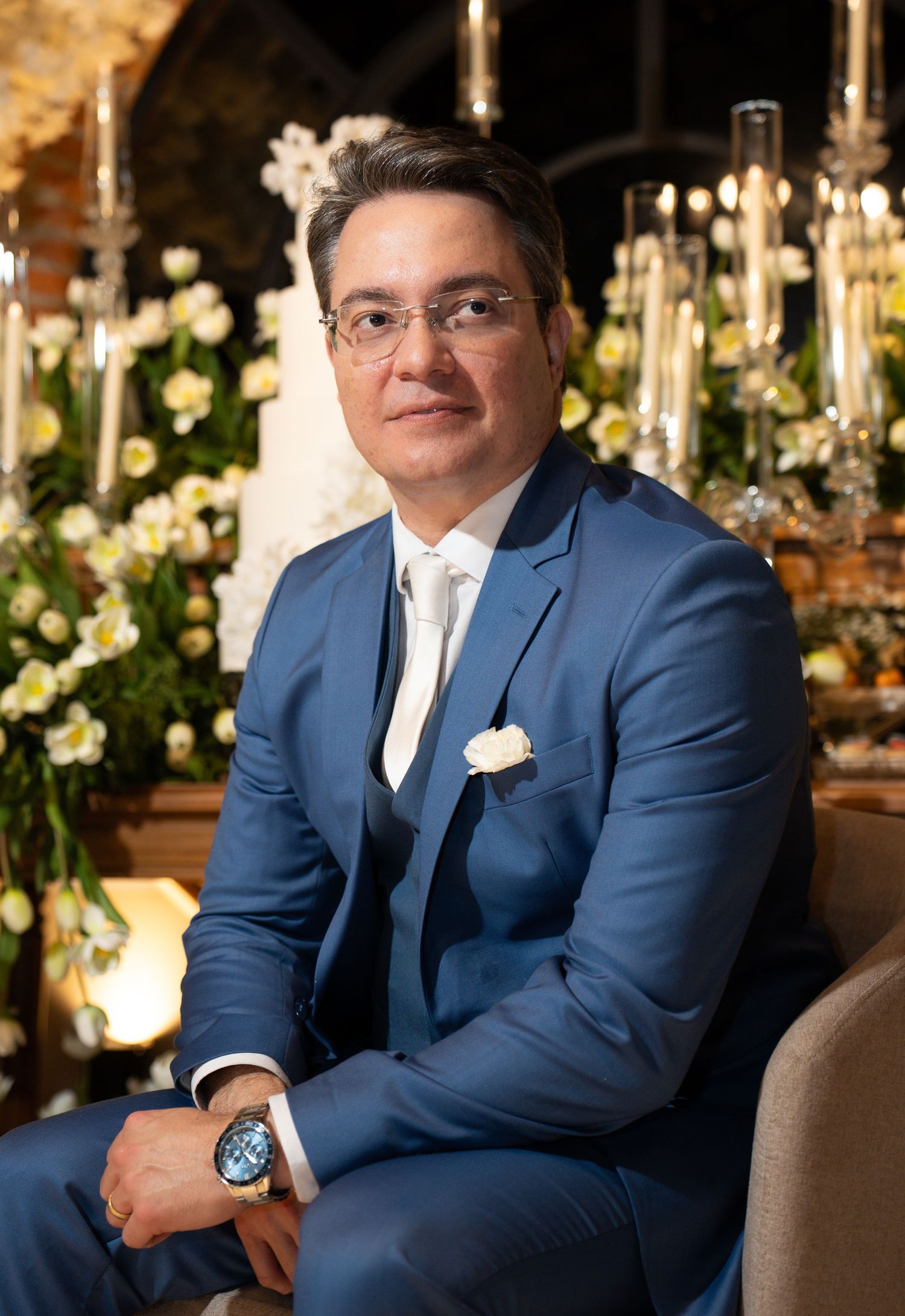 Elegant man in blue suit with white boutonniere seated at floral wedding reception backdrop.