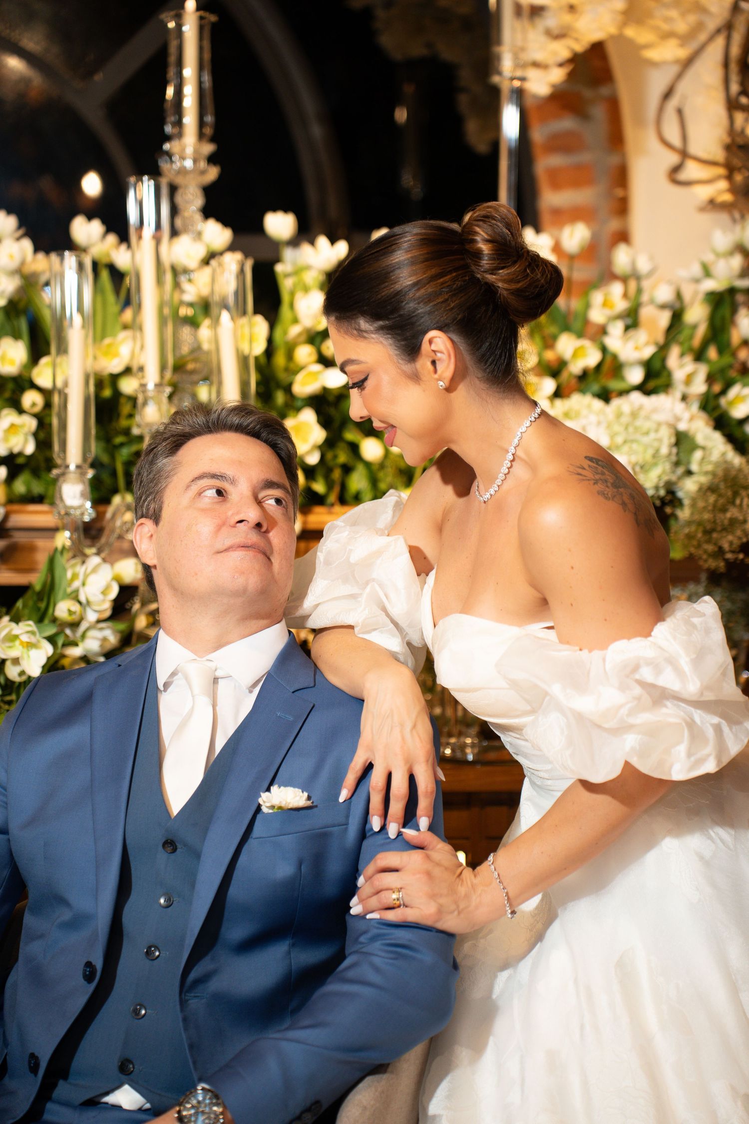 Bride in white off-shoulder gown and groom in blue suit share a romantic moment surrounded by white floral arrangements.