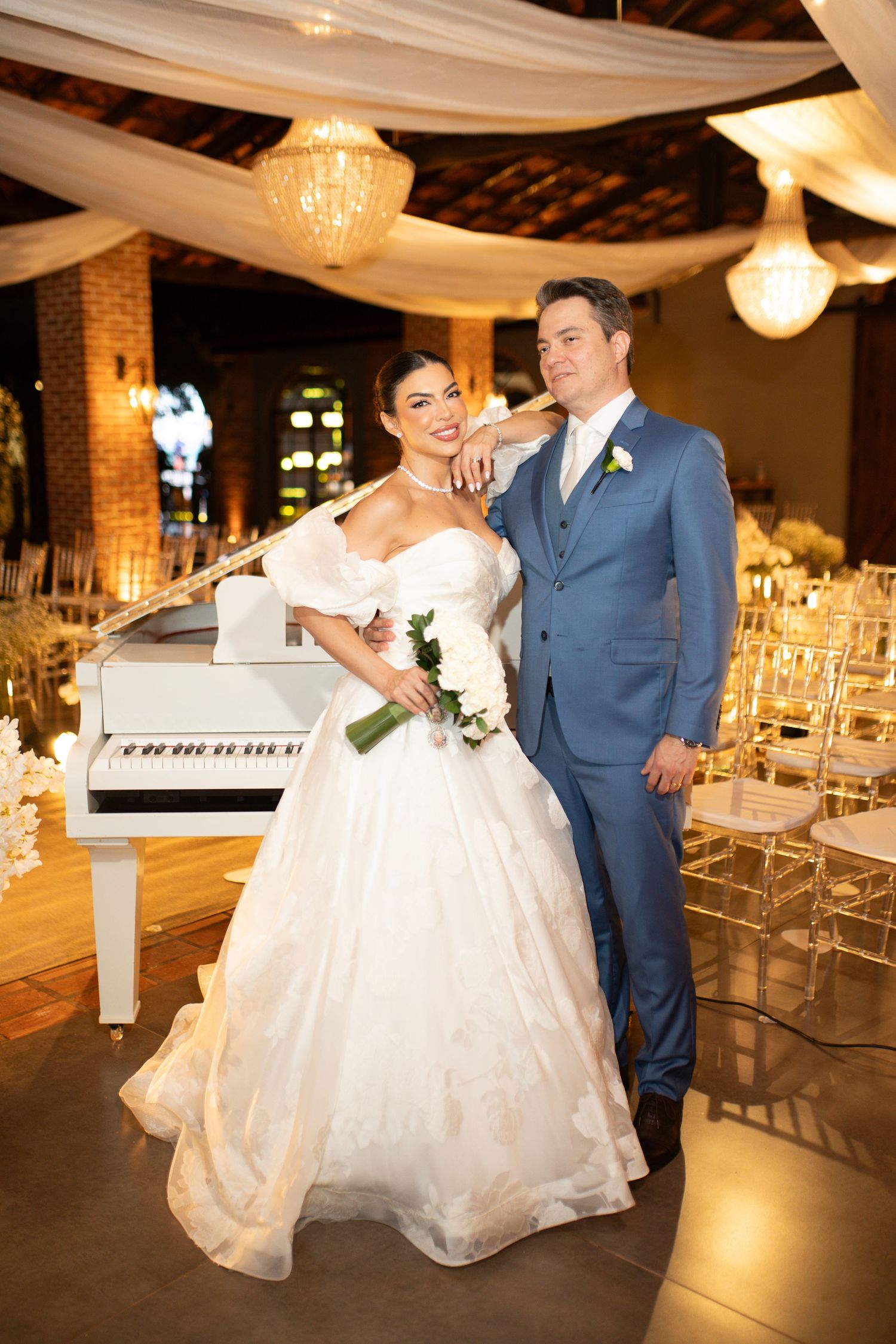 Bride in white ballgown and groom in blue suit pose together near a white grand piano at elegant wedding venue.
