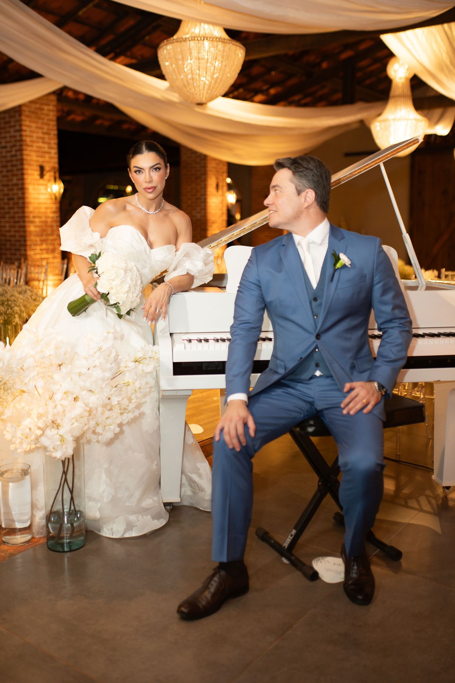 Bride in white gown holds bouquet beside groom in blue suit seated at white grand piano in elegant wedding venue.