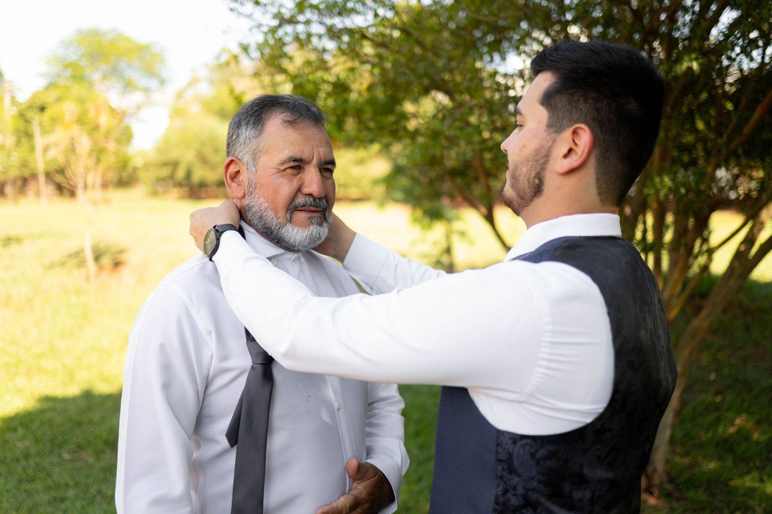 A young man in a vest helps an older gray-bearded man adjust his tie outdoors at a wedding.
