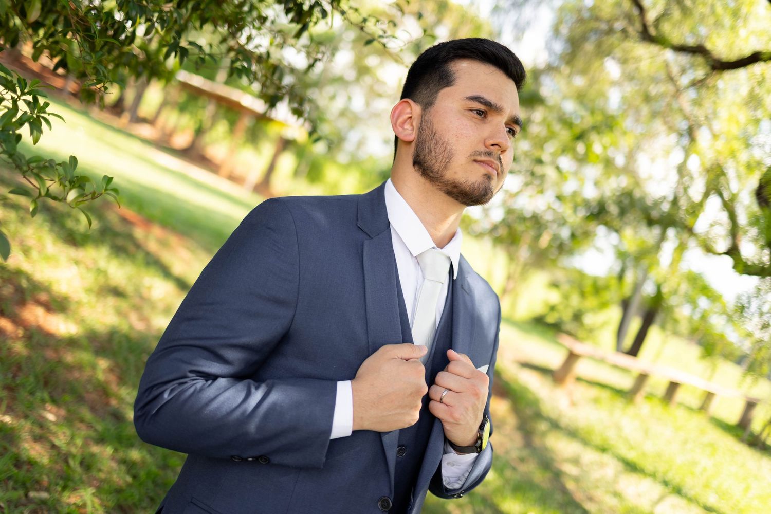 Young man in navy blue suit adjusting jacket lapel outdoors in green park setting.
