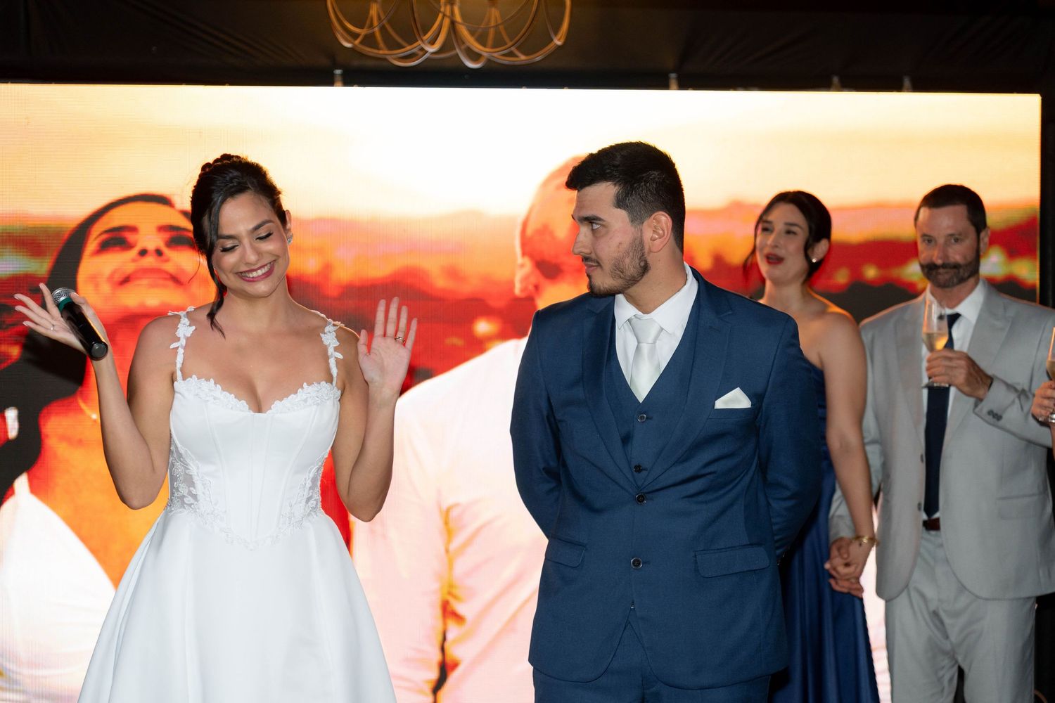 A bride in a white gown and groom in a navy suit smile during their wedding reception with guests in the background.