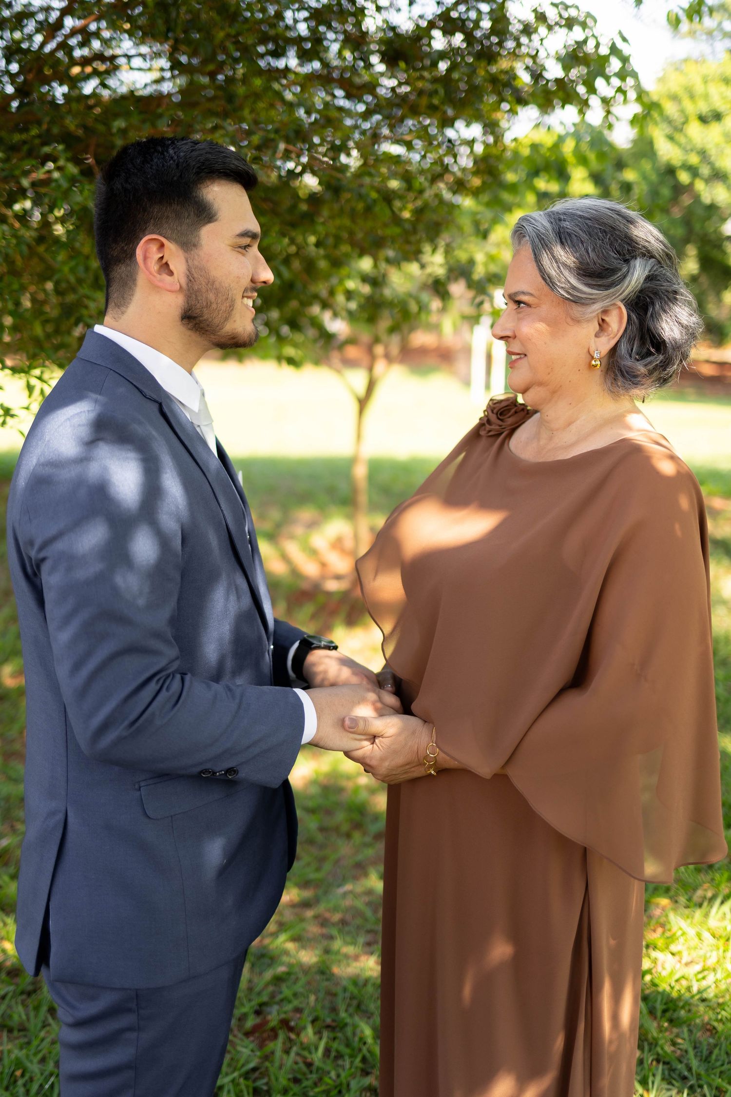 Young man in blue suit holds hands with elegant older woman in brown dress, smiling outdoors in a garden.