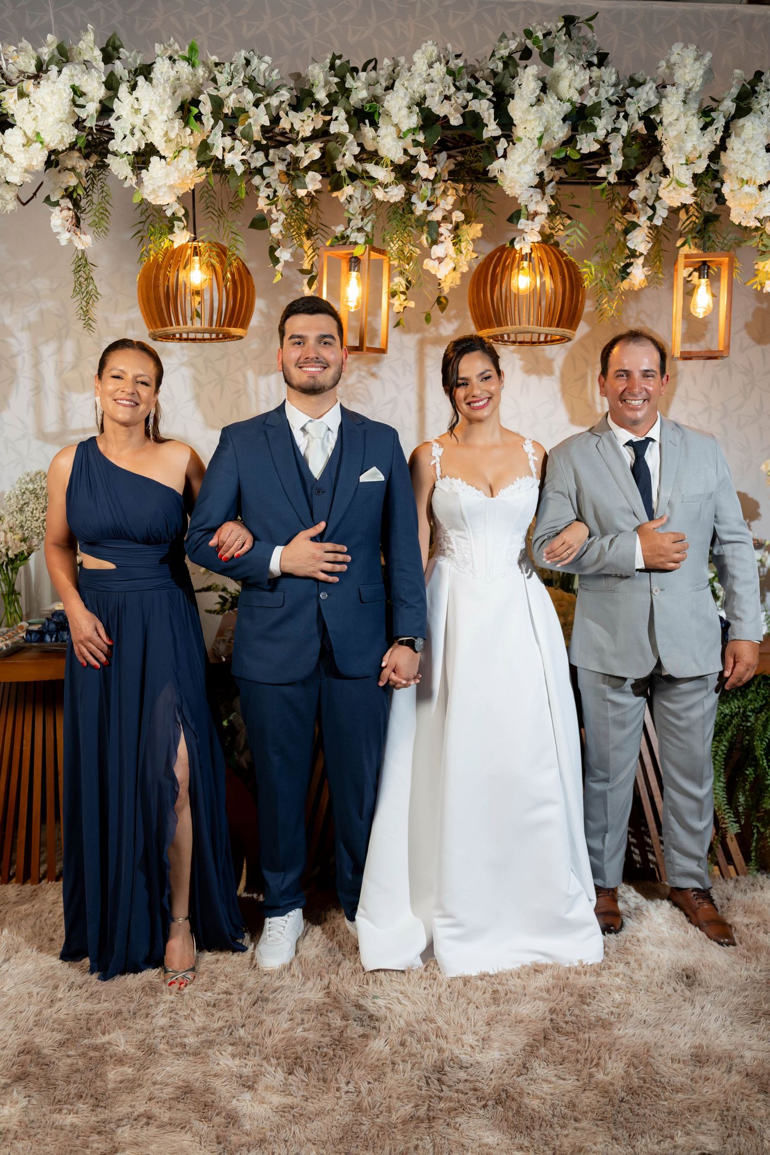 A bride and groom pose with two guests in elegant attire beneath floral decorations and warm pendant lights.