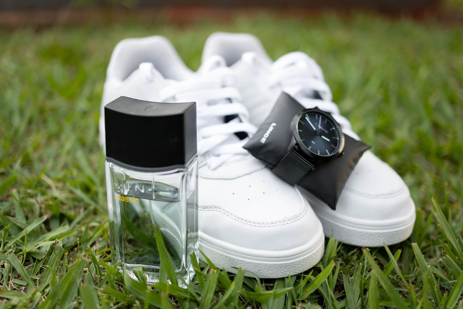White sneakers, a black wristwatch, and a glass cologne bottle arranged on green grass.