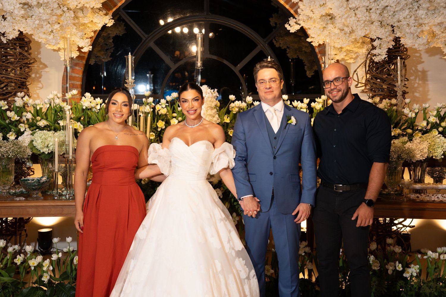 Elegant wedding ceremony with bride in white gown, groom in blue suit, and two guests posing amid floral decorations.