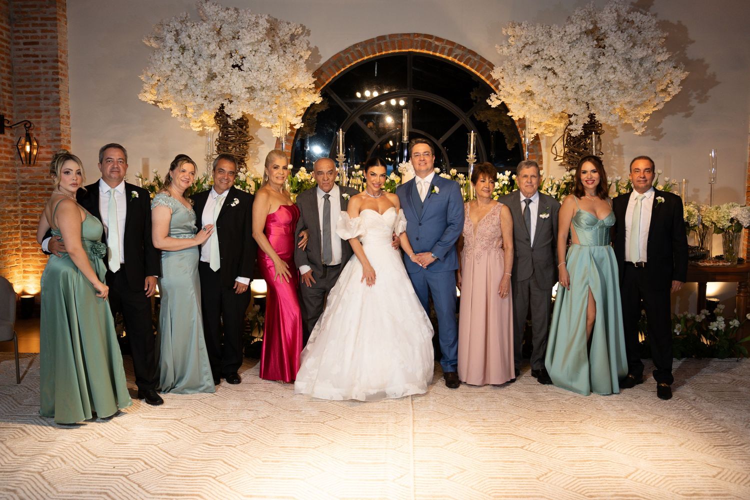 Wedding party poses together in elegant venue with floral arch, bride in white gown surrounded by guests in formal attire.