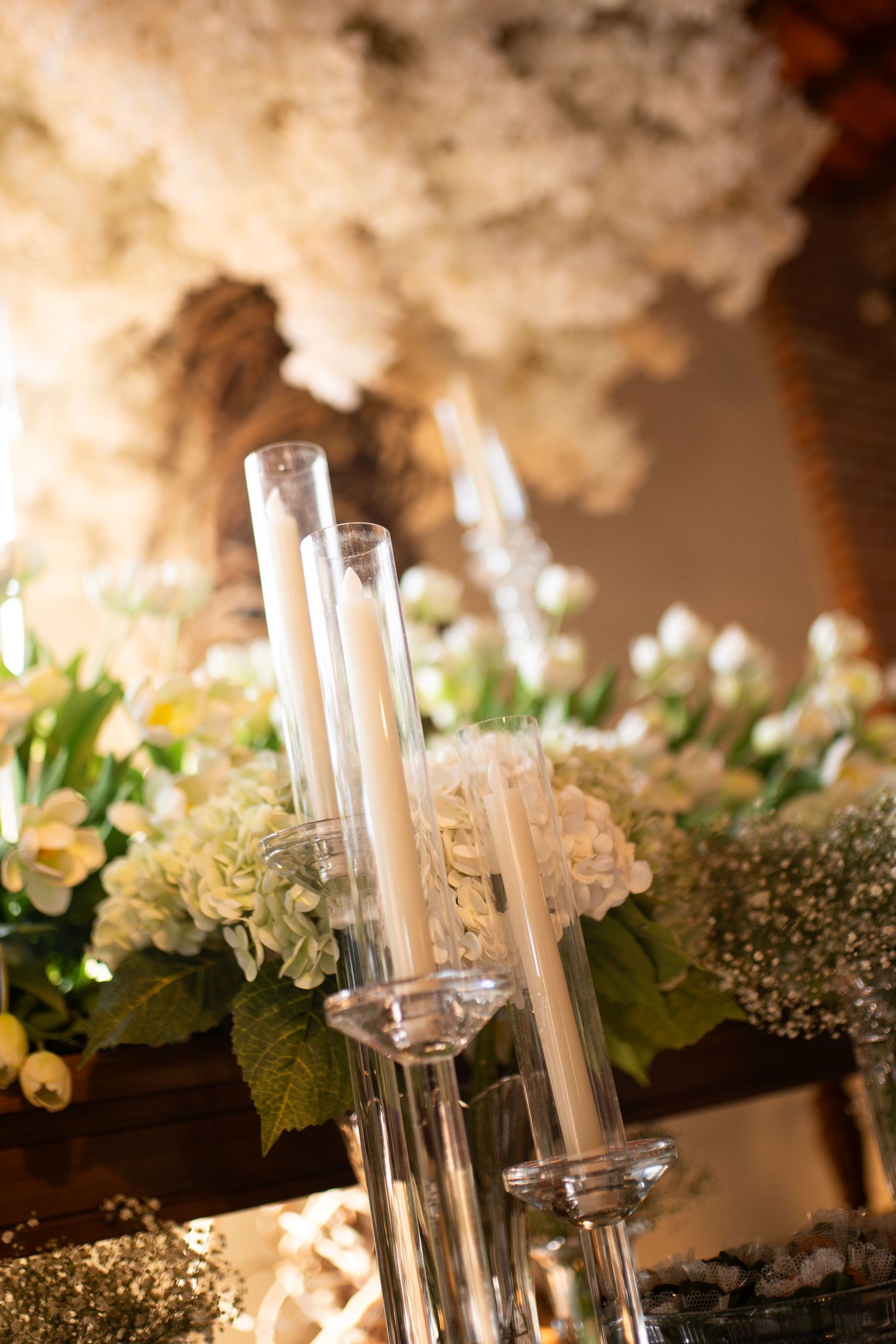 Elegant tall glass candle holders with white taper candles surrounded by lush white floral arrangements at a wedding.