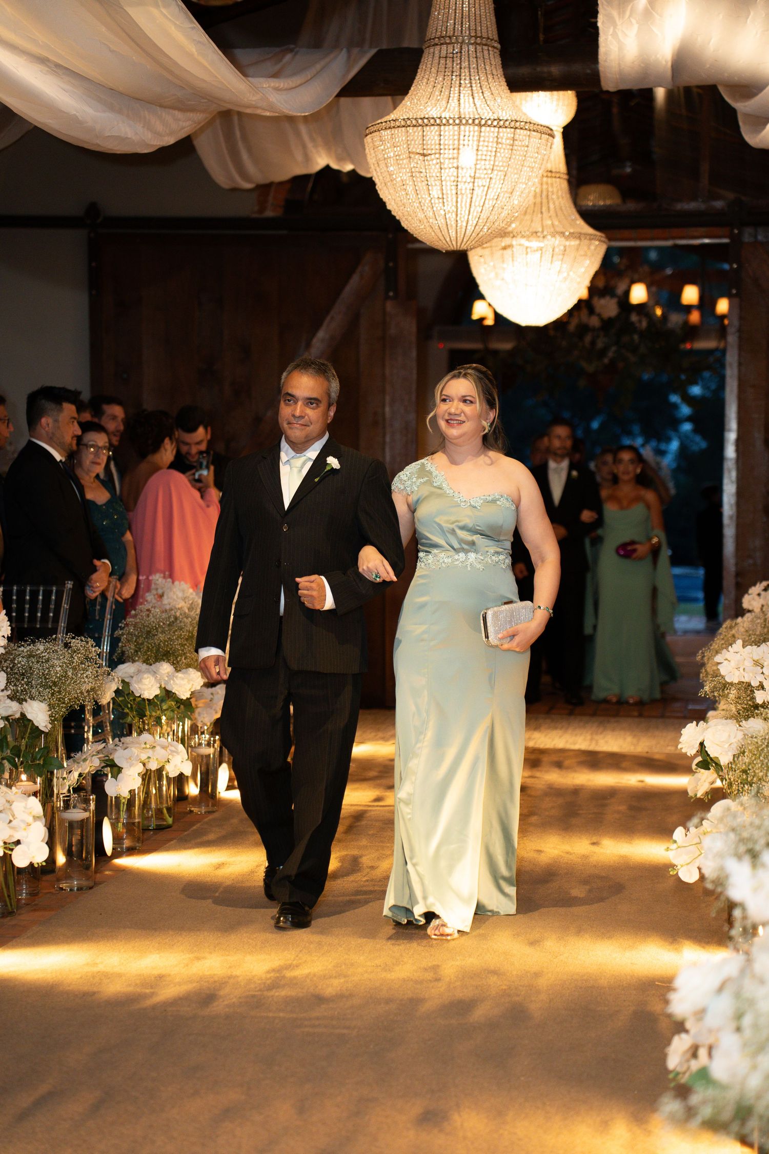Elegant wedding ceremony with couple walking down aisle under crystal chandeliers, woman in mint gown, man in tuxedo.