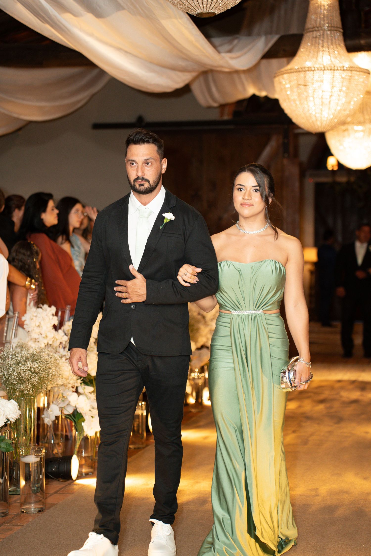 Elegant couple walking down wedding aisle, man in black suit, woman in green strapless gown, chandelier backdrop.