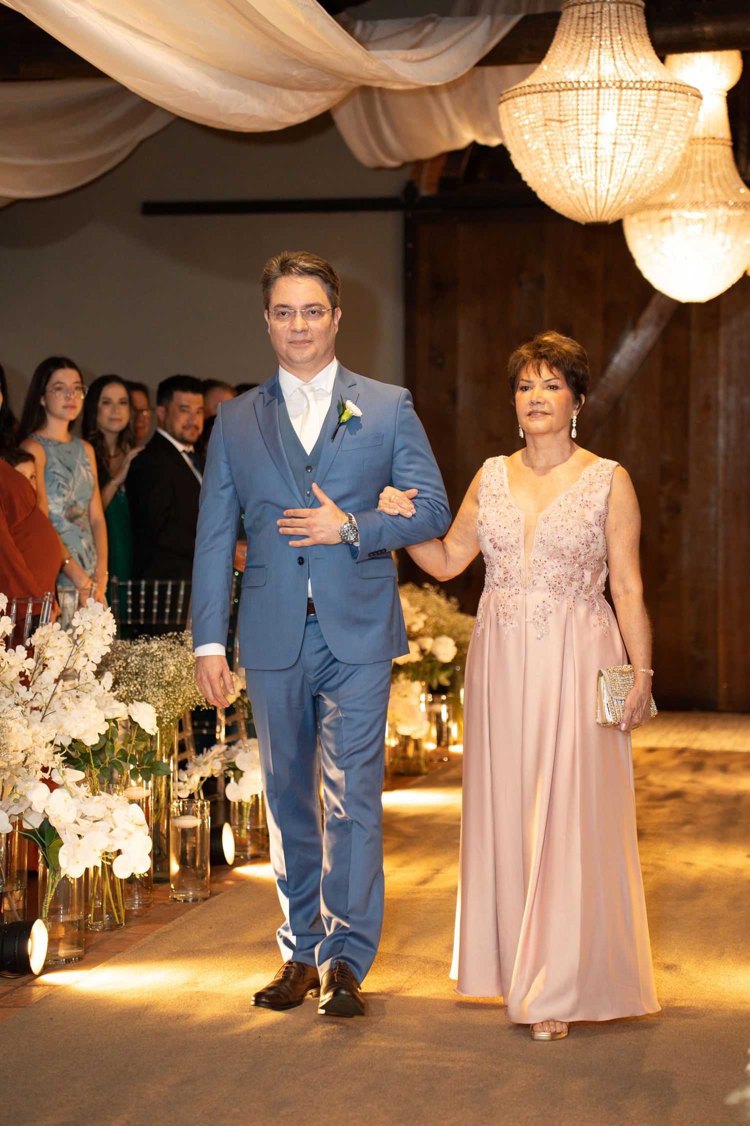 Groom in blue suit walks down wedding aisle with elegantly dressed woman in pink gown at decorated venue.
