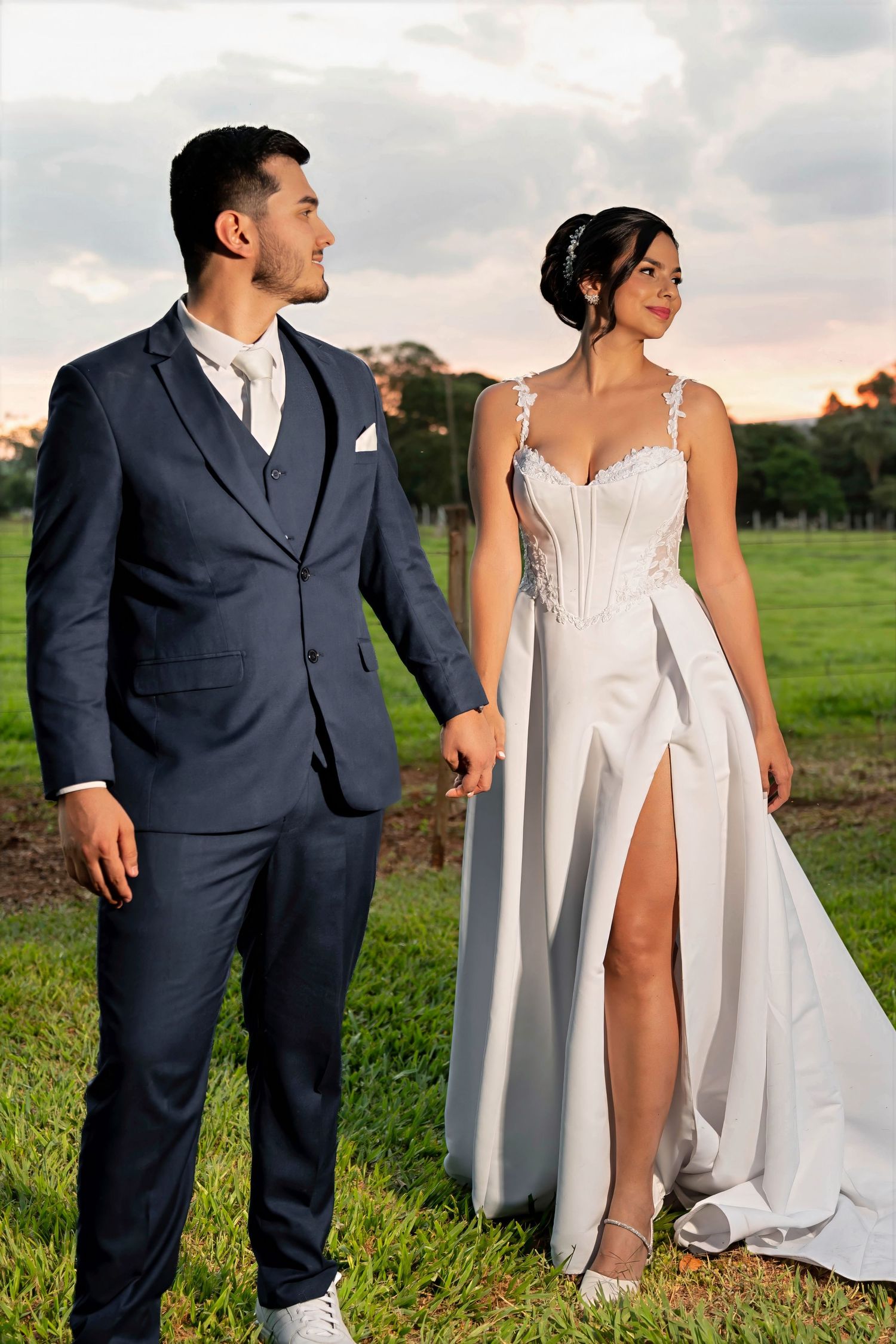 A couple in wedding attire holds hands outdoors, groom in navy suit, bride in white gown with lace bodice and slit skirt.