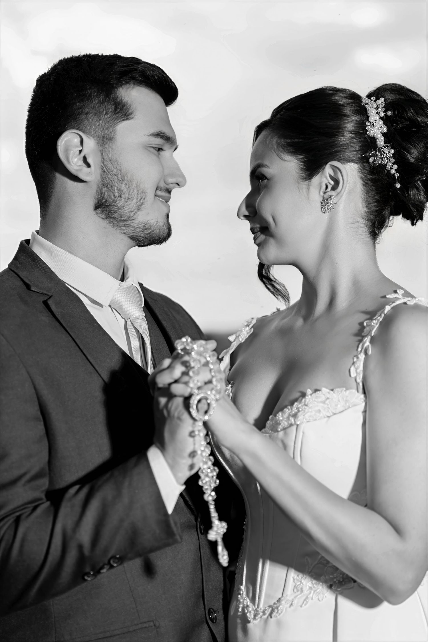 Black and white photo of a smiling bride and groom in wedding attire gazing at each other while holding a rosary.