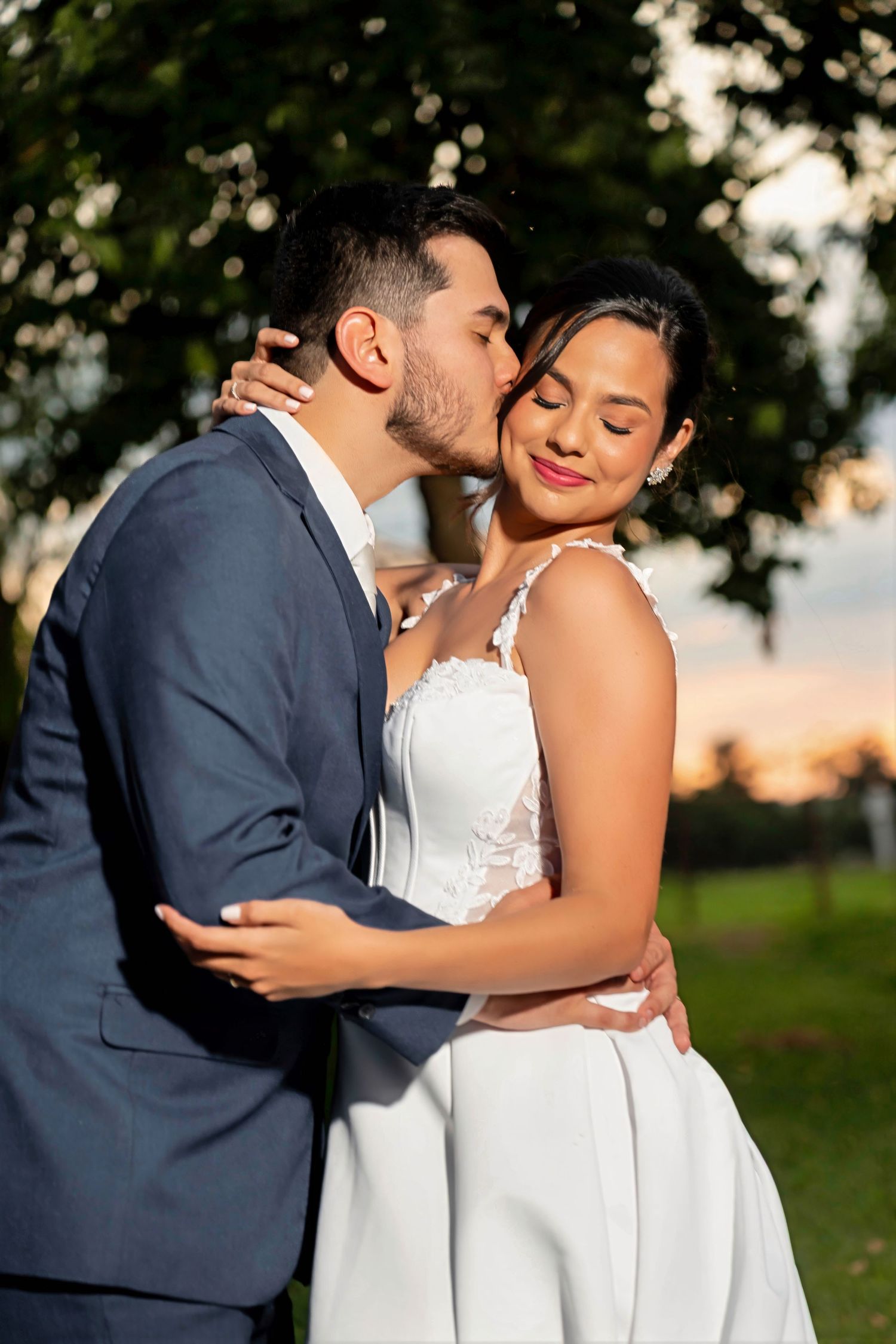 Groom in blue suit kisses bride in white lace wedding dress outdoors at sunset.