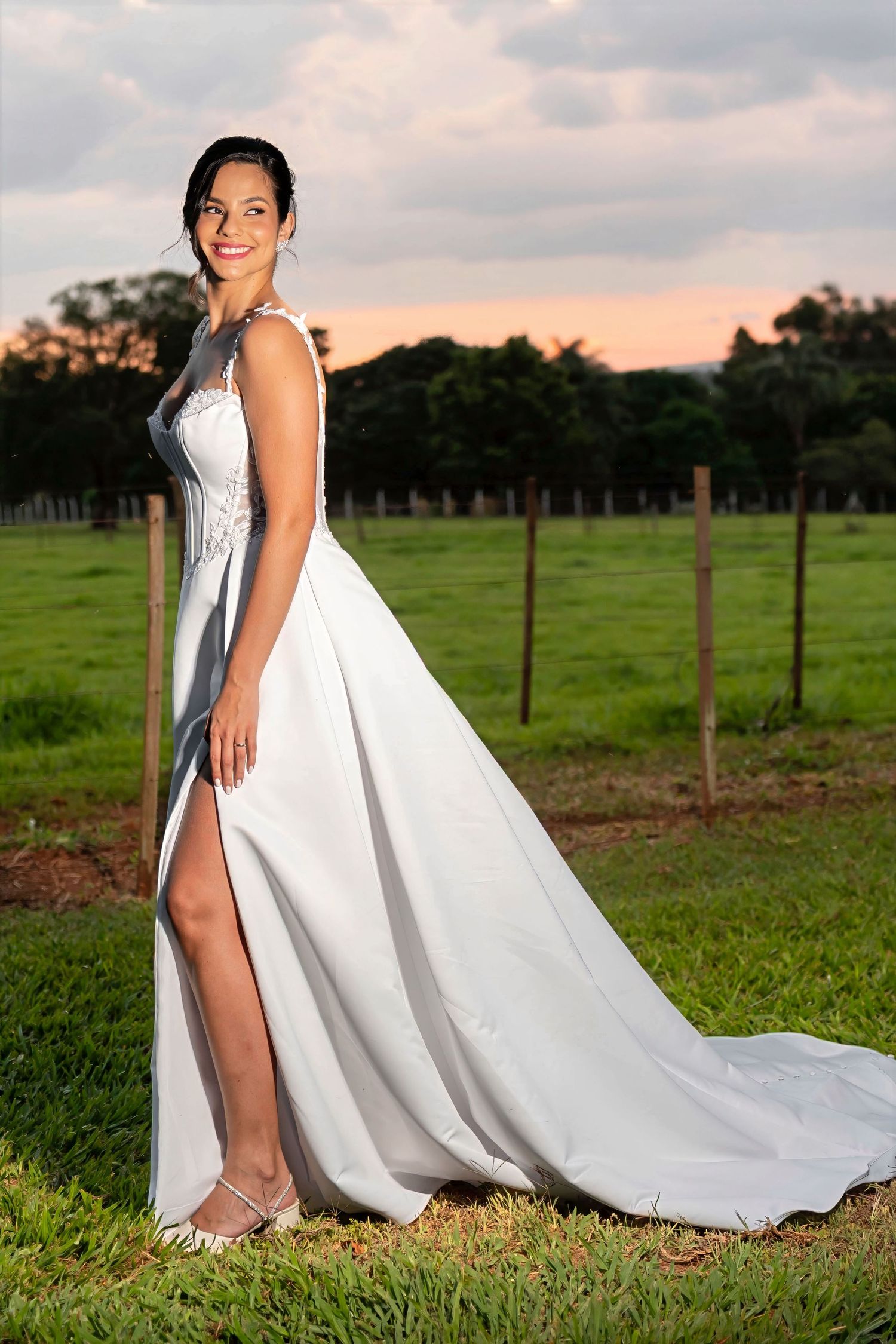 Smiling bride in white slit gown with lace bodice poses outdoors on green grass at sunset.