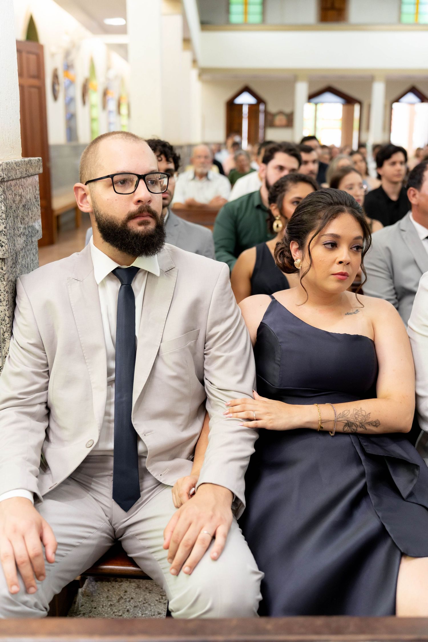 A bearded man in a gray suit and a woman in a navy dress sit together in church pews at a wedding ceremony.