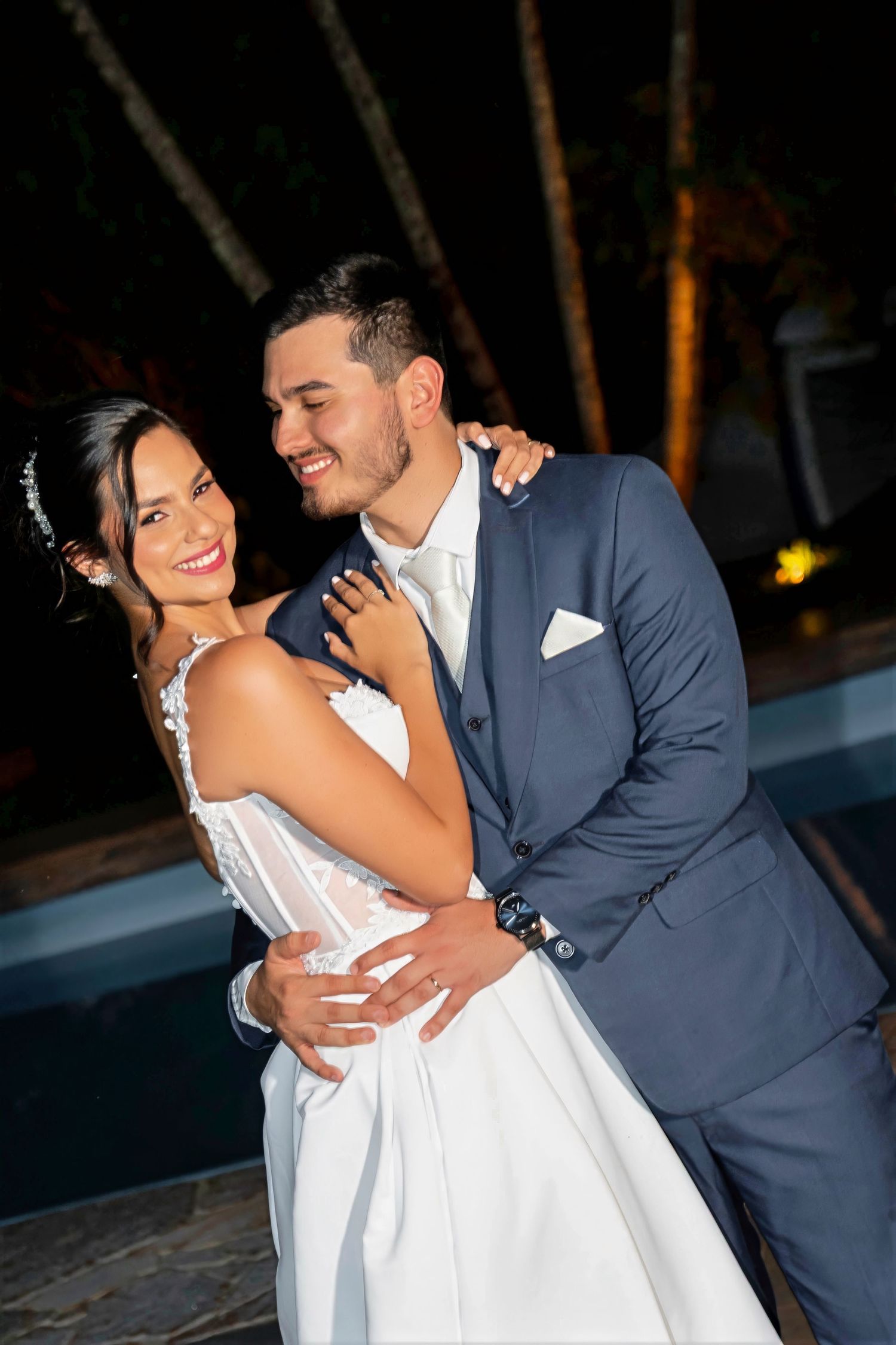A smiling bride in a white gown and groom in a navy suit share a romantic dance outdoors at night near a pool.