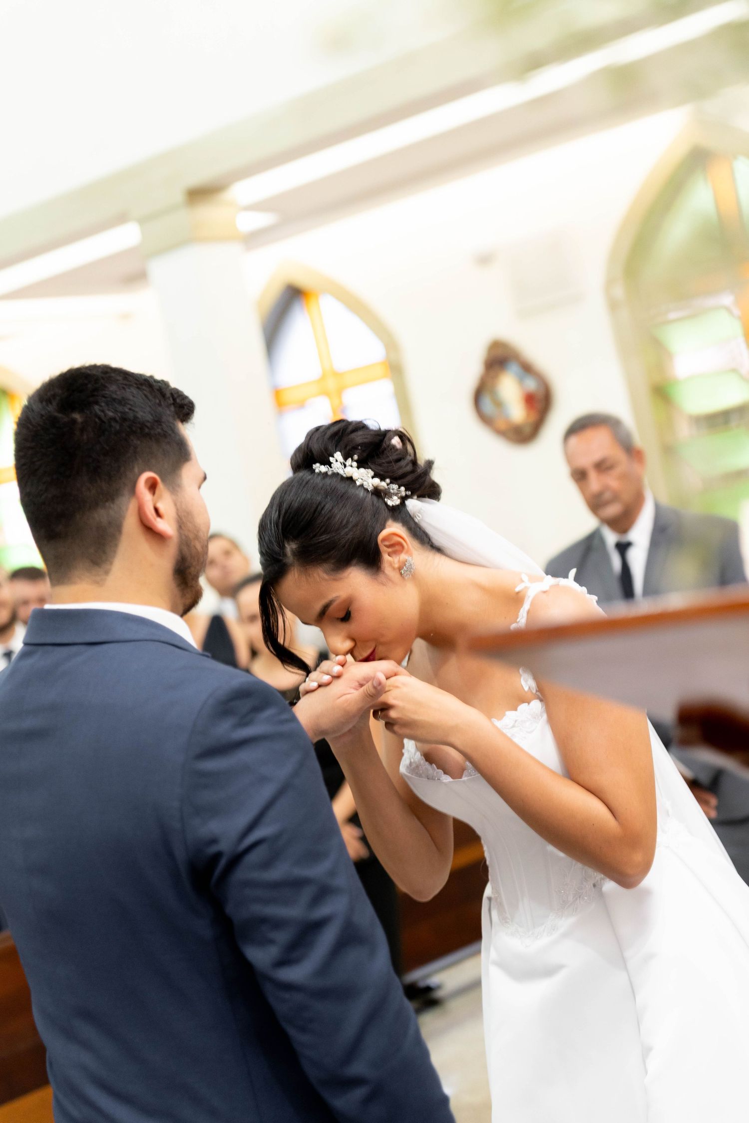 Bride kissing groom's hand during wedding ceremony in church, wearing white gown and floral hair accessory.