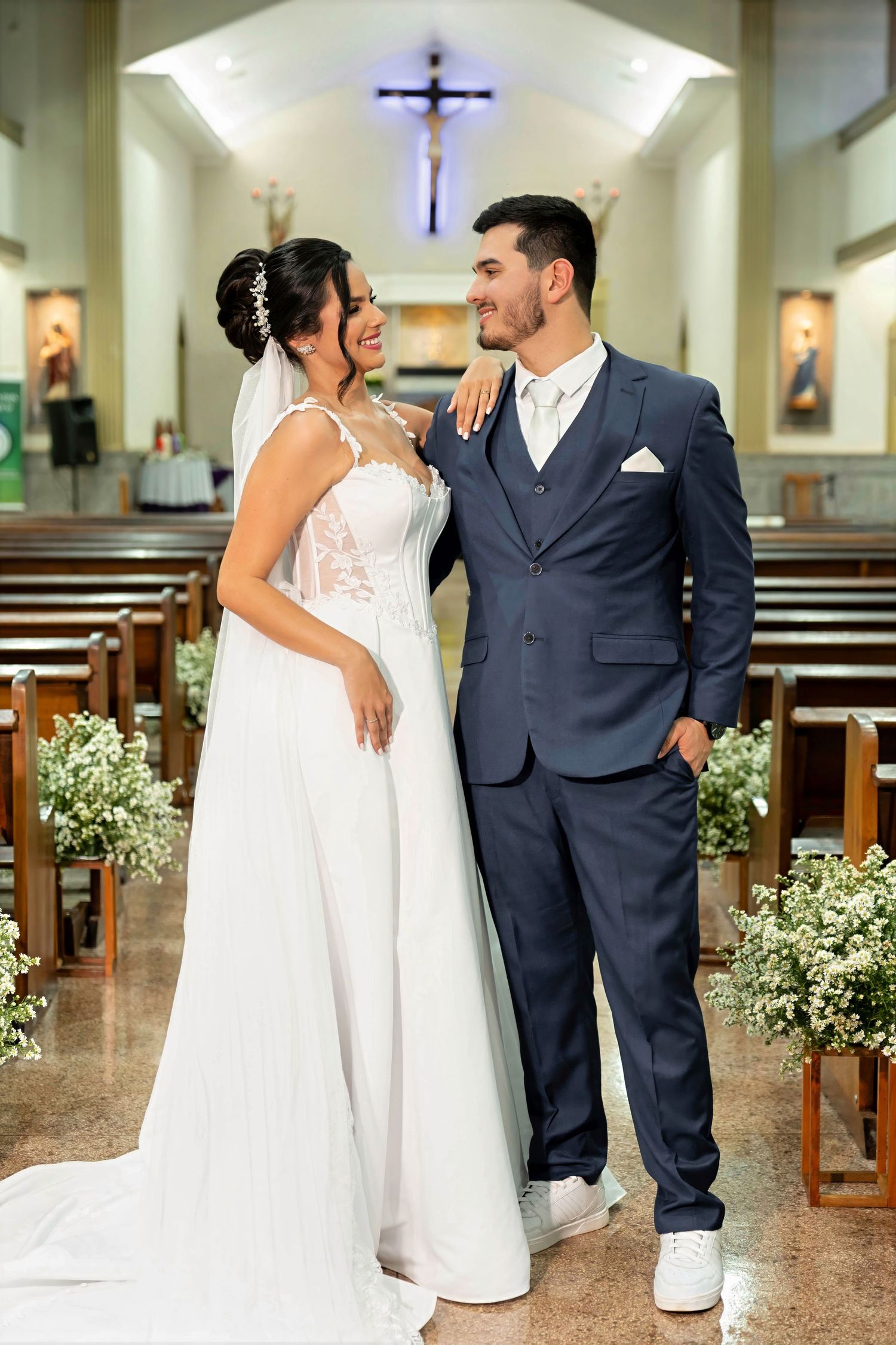 Bride and groom pose together in a church aisle, she in a white lace gown, he in a navy suit with white sneakers.