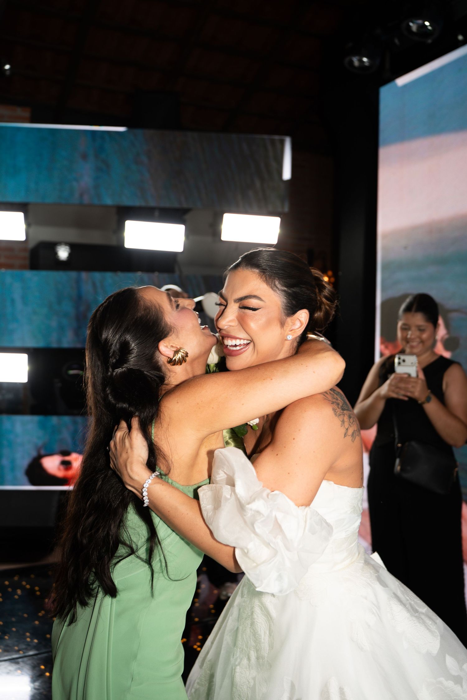 Two women in formal attire embrace warmly at an event, one in a white gown and one in mint green.