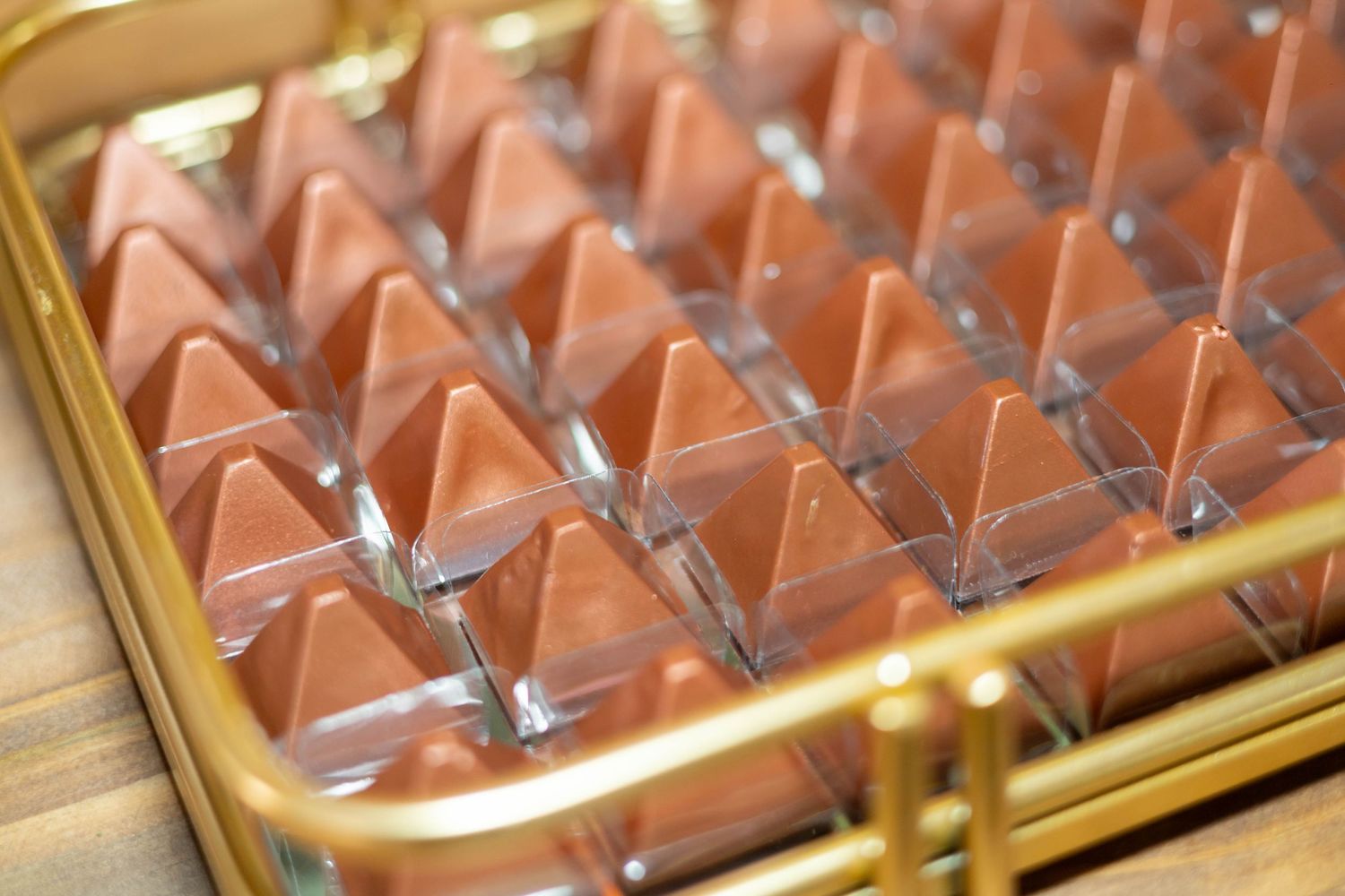 A gold tray filled with pyramid-shaped milk chocolates in clear plastic packaging on a wooden surface.