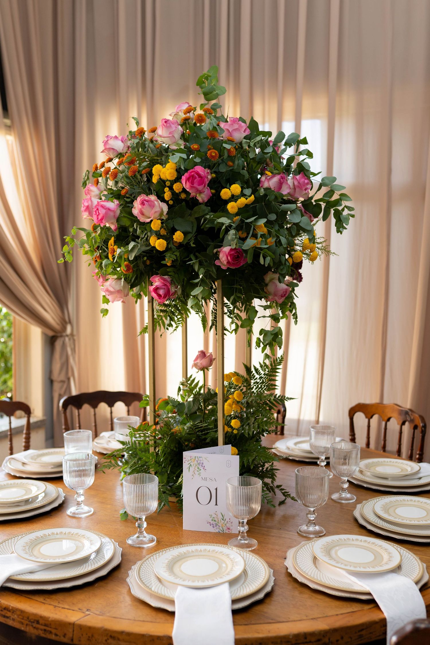 Elegant wedding table setting with tall floral centerpiece of pink roses and yellow blooms on a round wooden table.