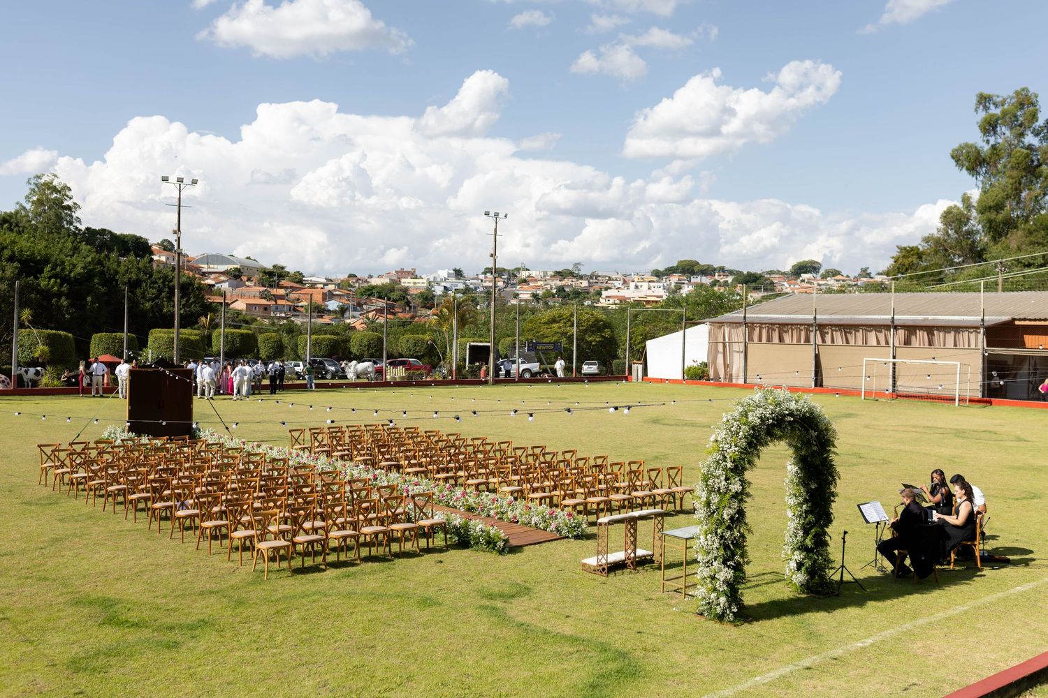 Outdoor wedding ceremony setup on green field with rows of chairs, floral arch, and city skyline backdrop.