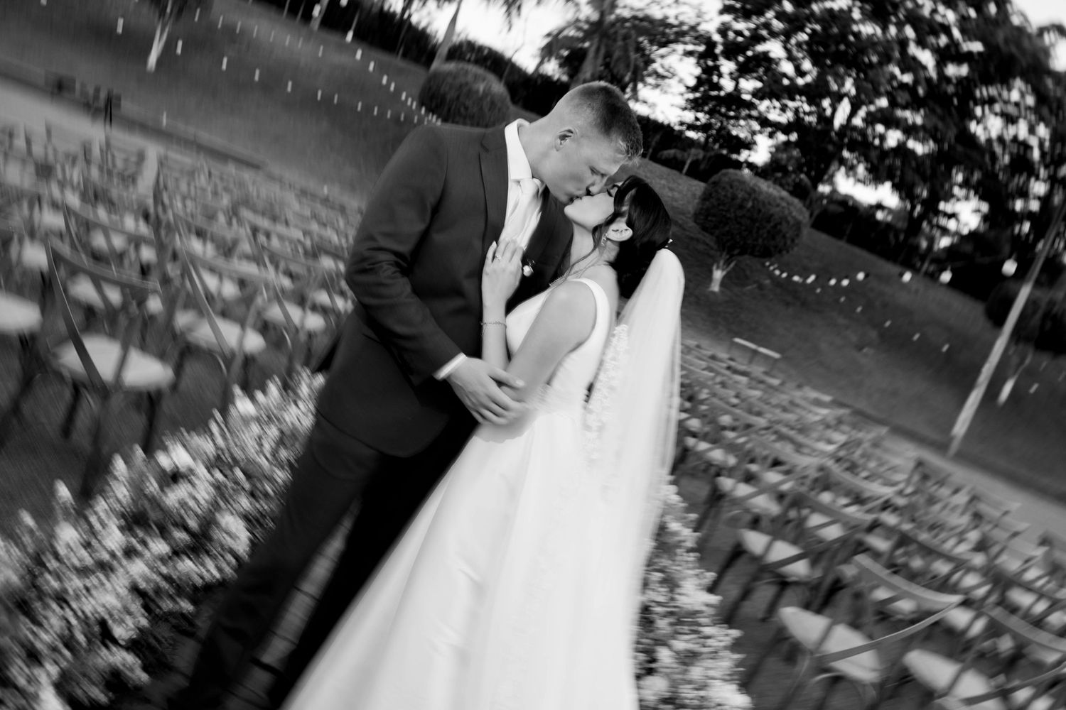 Black and white photo of a bride and groom sharing a romantic kiss outdoors at their wedding ceremony.