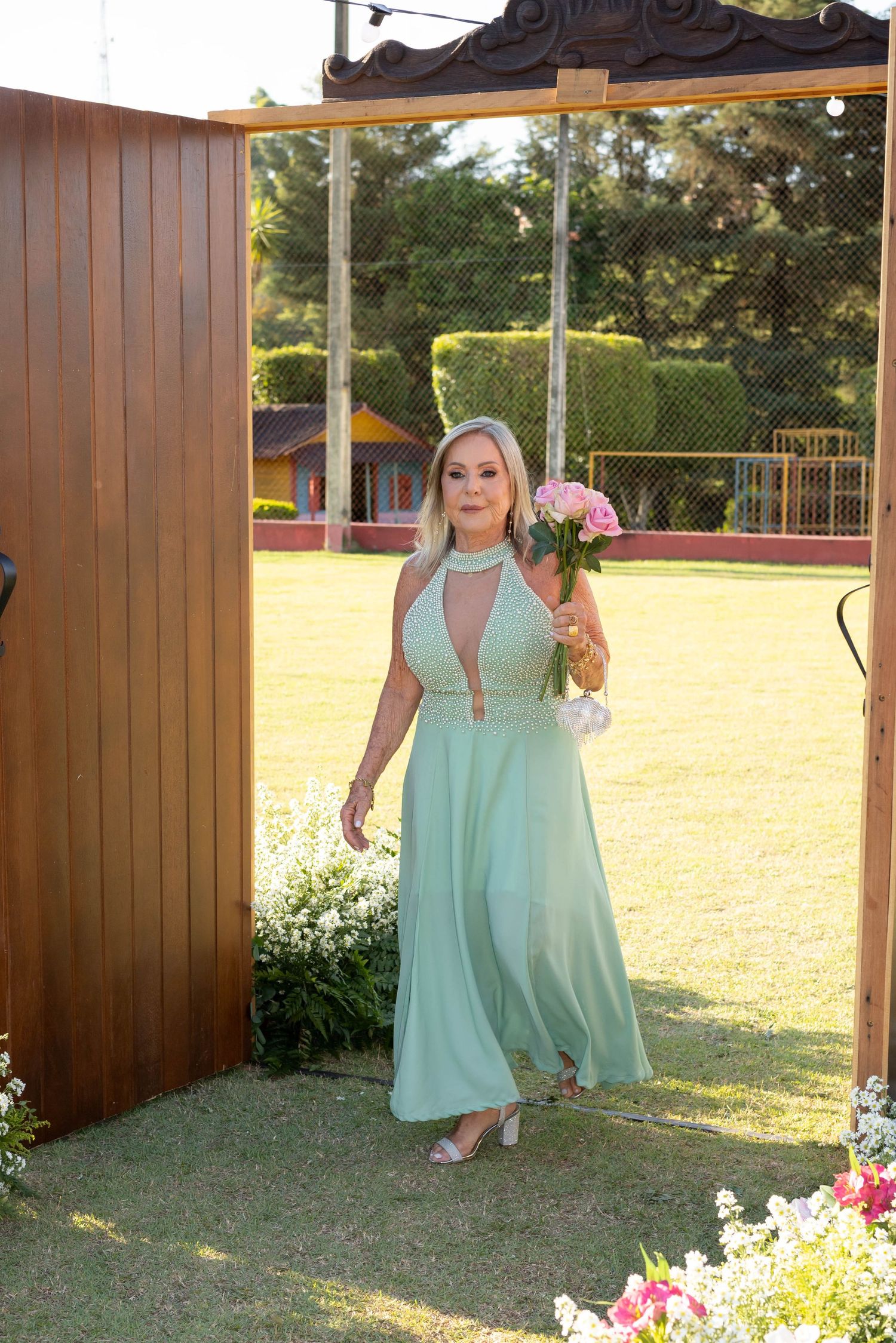 Elegant woman in mint green halter gown holding pink bouquet stands in wooden archway at outdoor wedding venue.