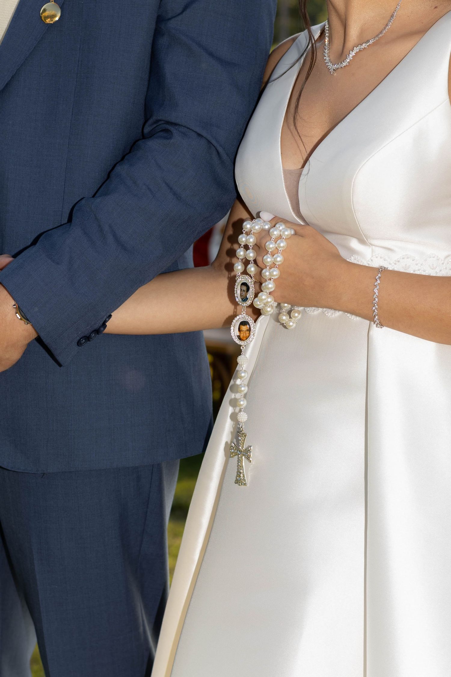Bride and groom holding a pearl rosary with cross pendant, wearing formal wedding attire at a ceremony.