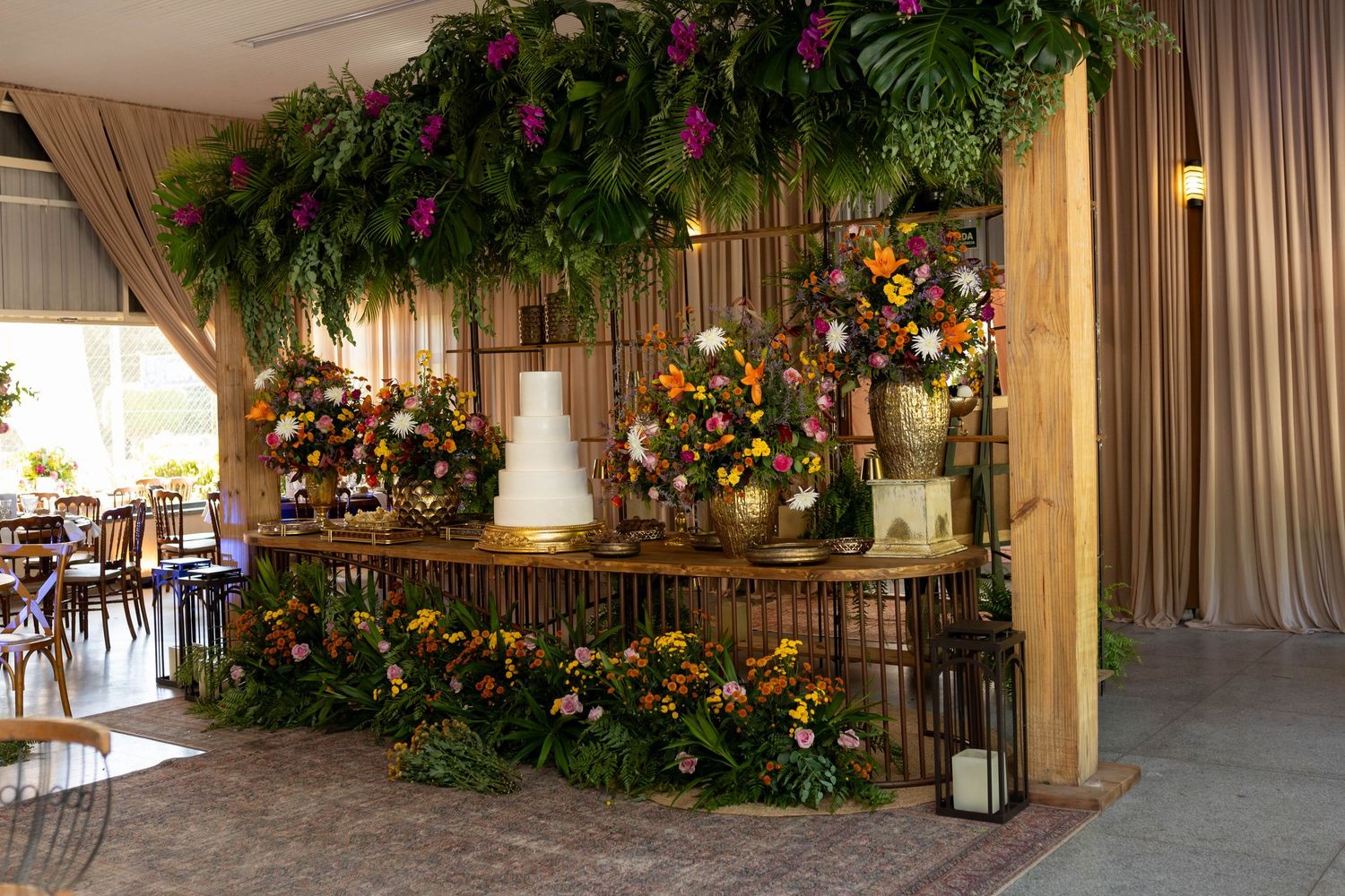 Elegant wedding cake table adorned with lush tropical greenery, colorful floral arrangements, and golden accents in a rustic venue.