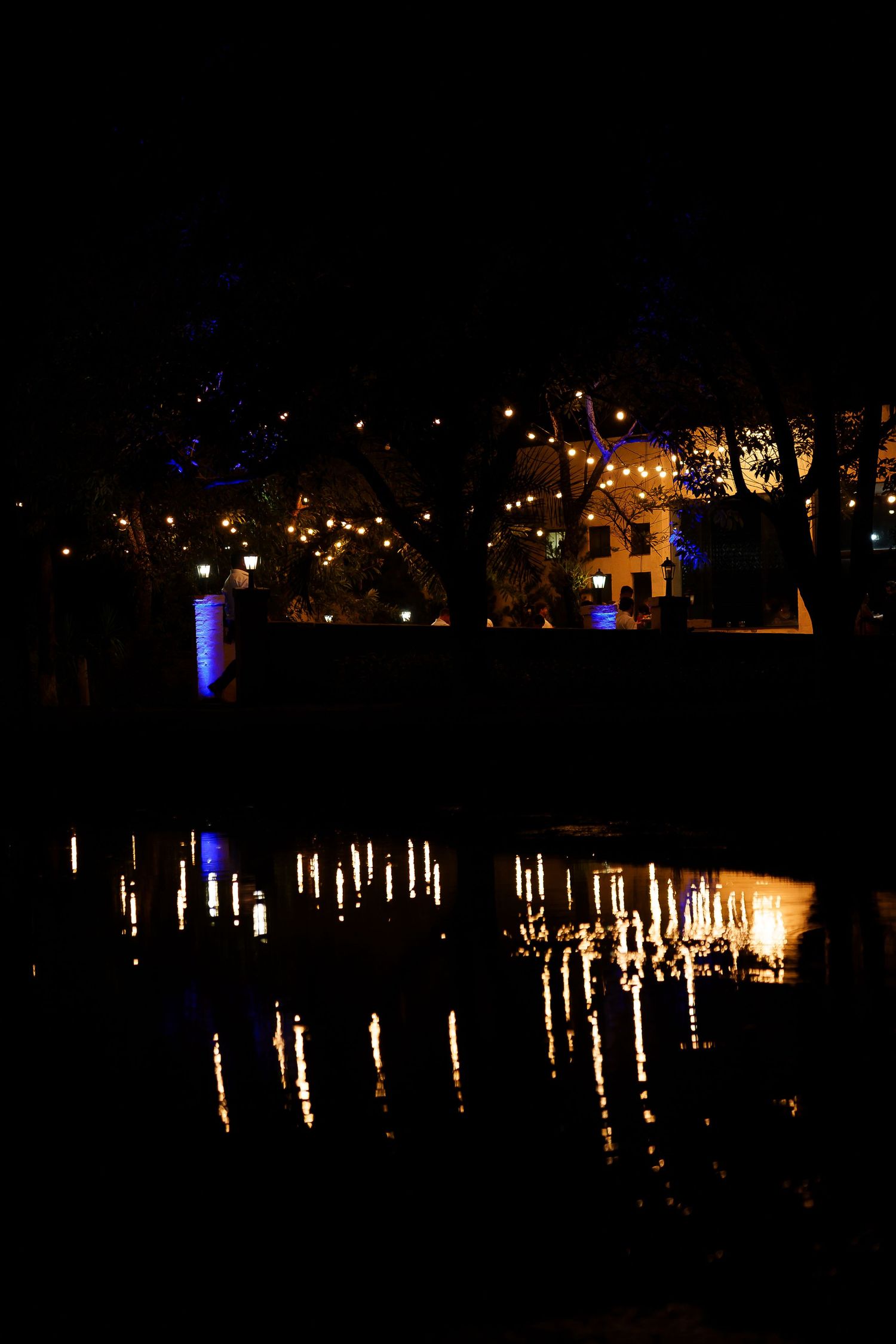 Night scene with glowing lights reflecting on calm water beside illuminated buildings and trees.