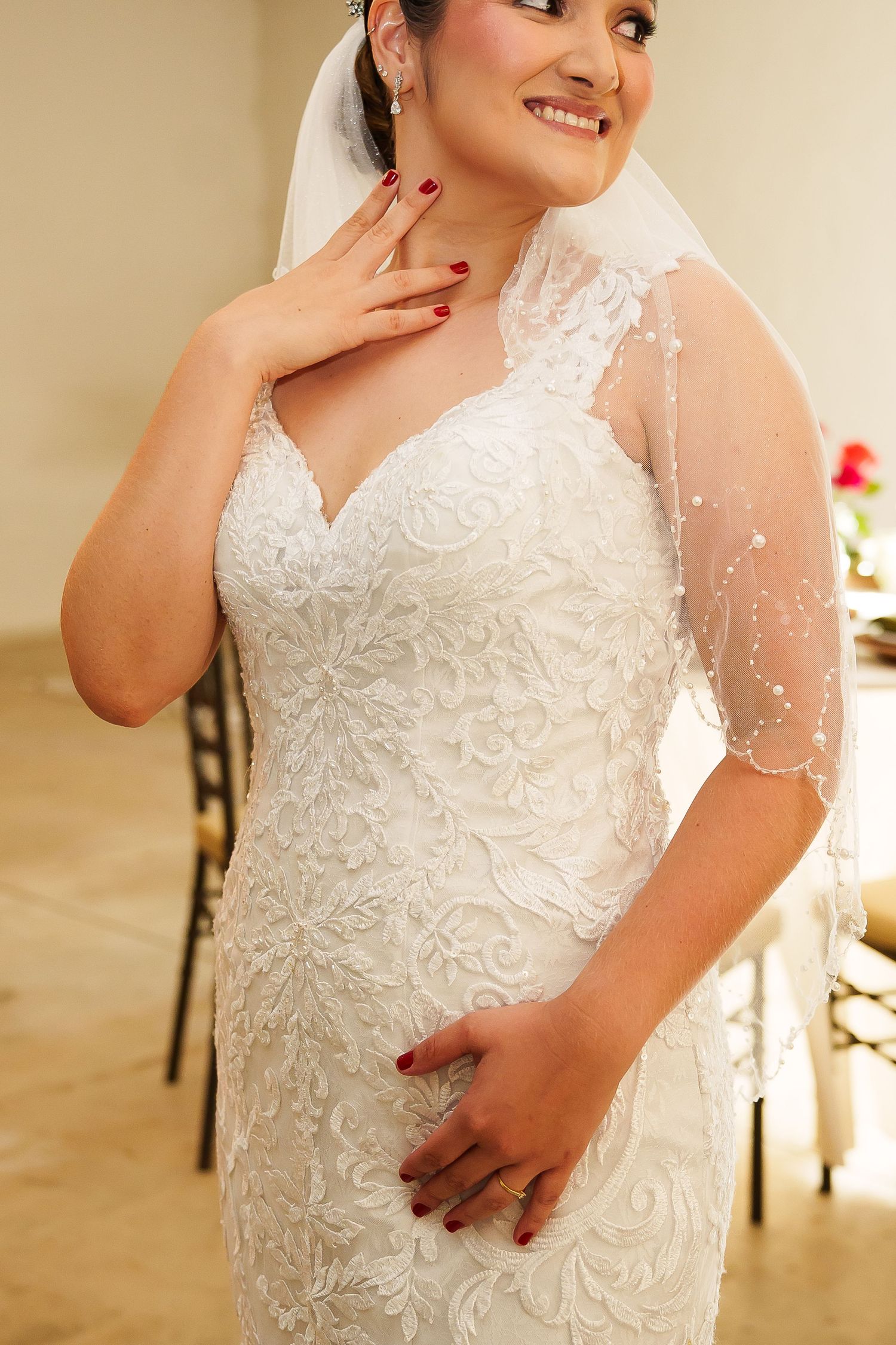 Smiling bride in white lace mermaid wedding gown with sheer veil, posing elegantly with hand on chest.