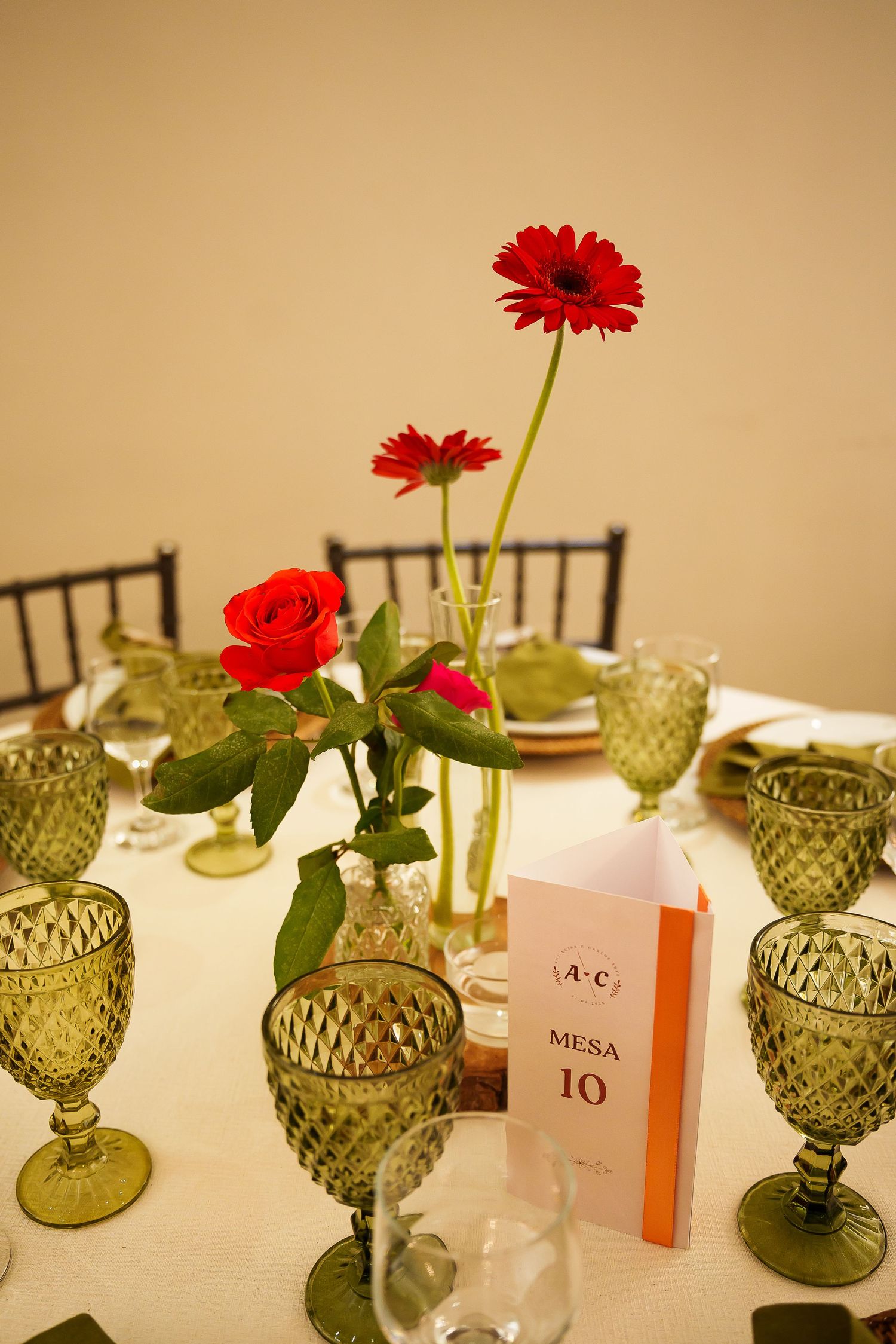 Elegant wedding table setting with red roses, gerbera daisies, green diamond-cut glasses, and a Mesa 10 table number card.