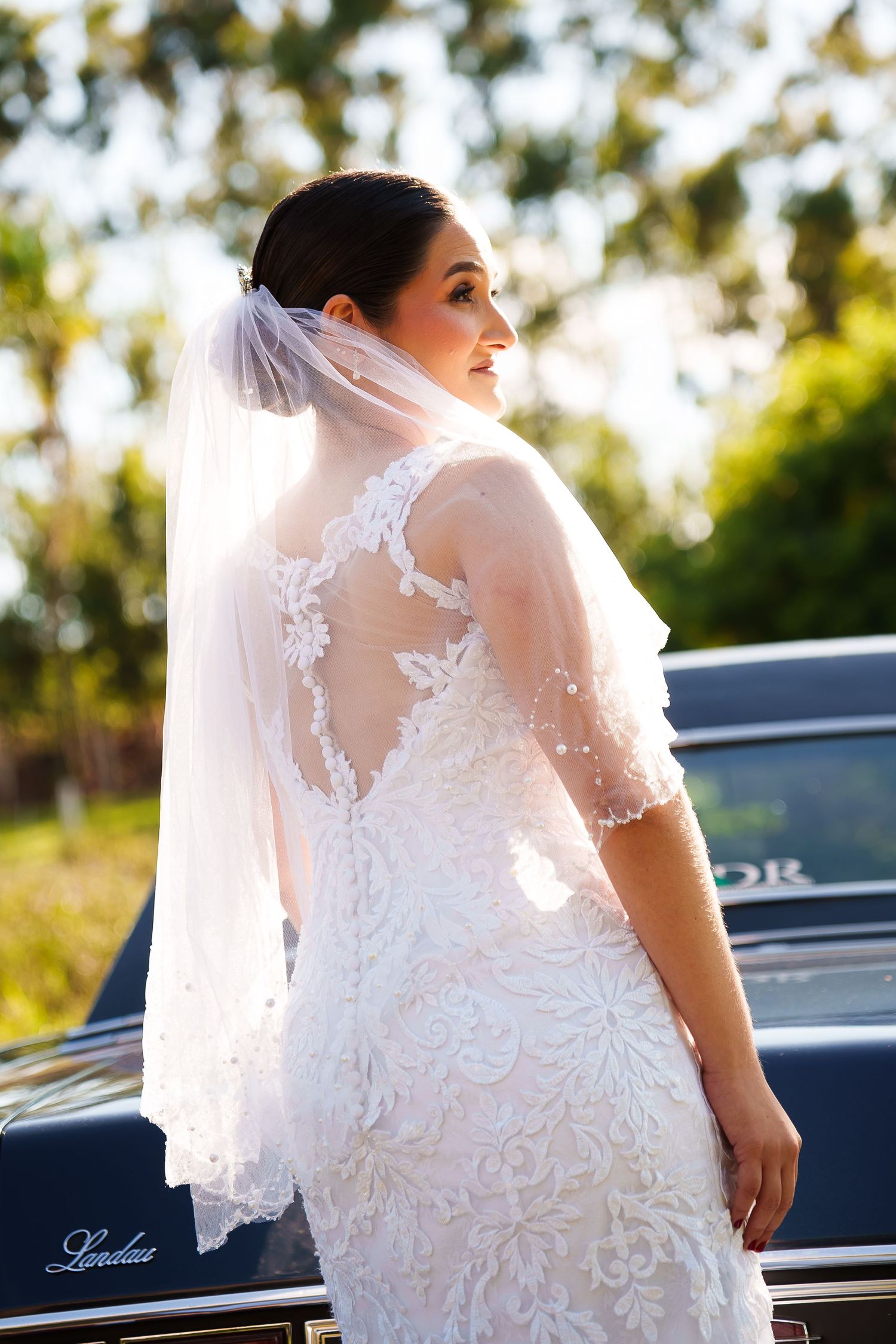 Bride in lace wedding dress and veil poses beside a classic black car outdoors with green trees in background.
