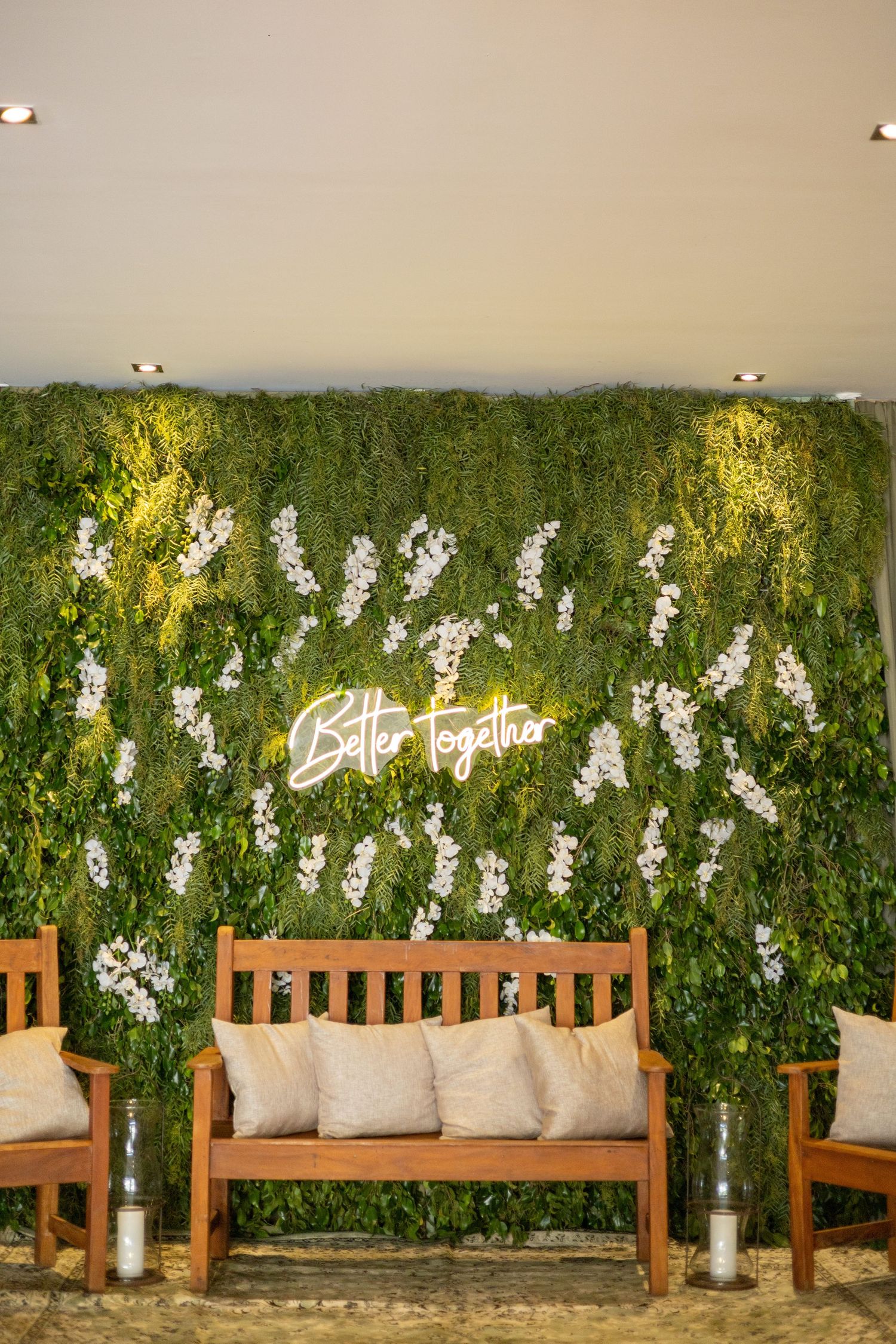 Green living wall with white orchids, neon 'Better Together' sign, wooden bench with cushions and candles.