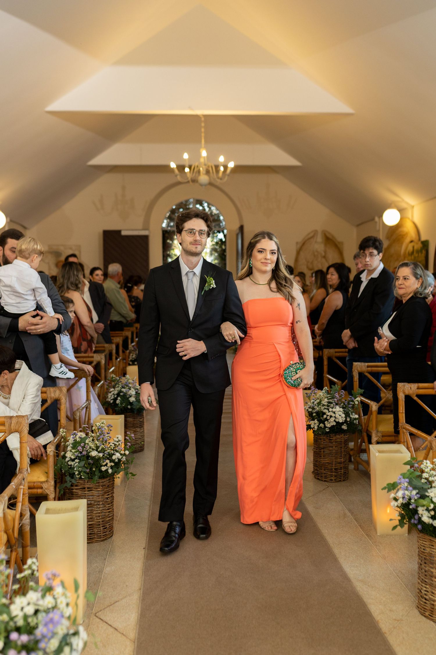 A couple walks down the aisle at a wedding, woman in orange gown, man in dark suit, chapel setting.