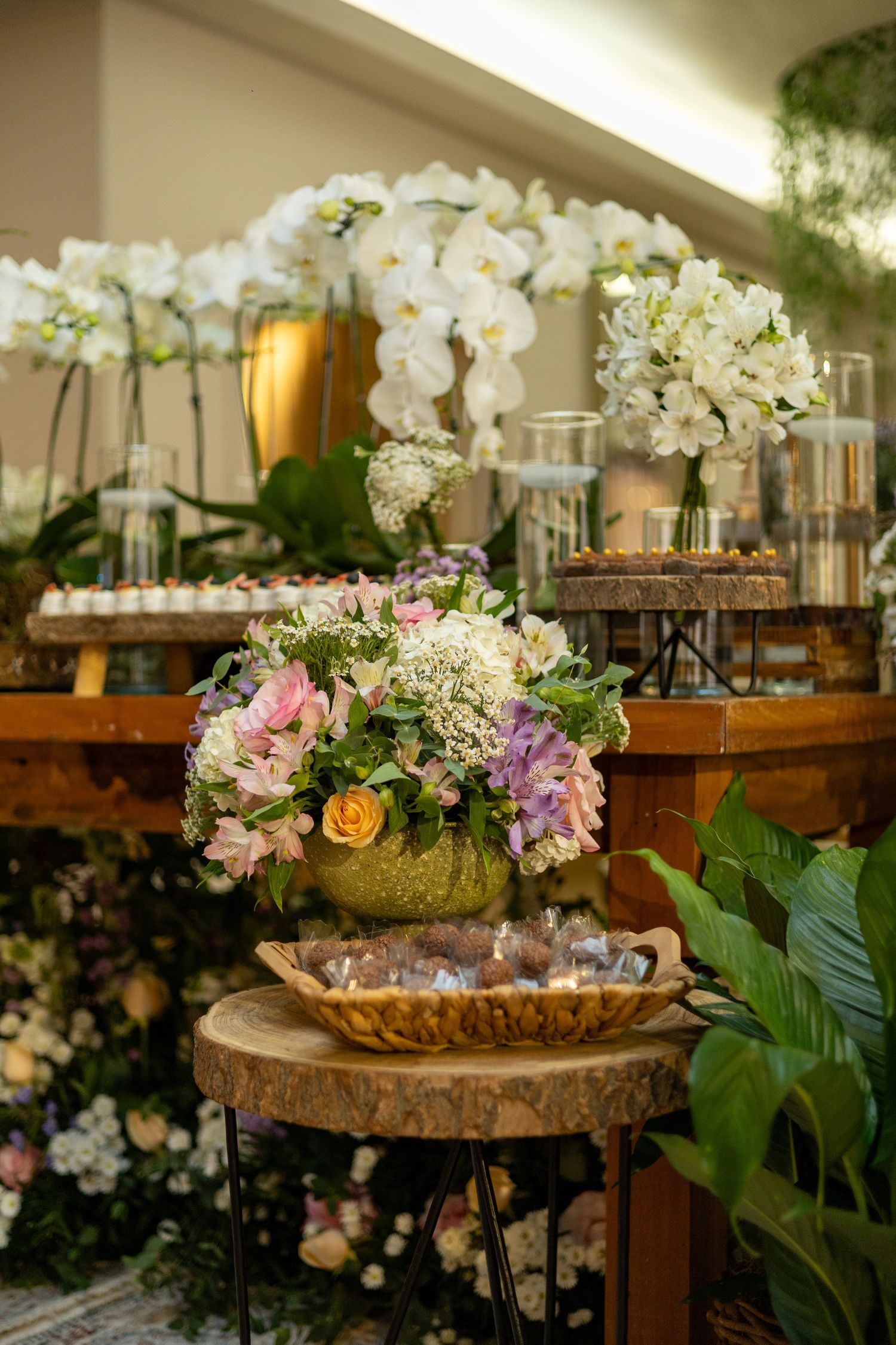 Elegant wedding dessert table with white orchids, mixed floral arrangements, and sweet treats on rustic wooden stands.