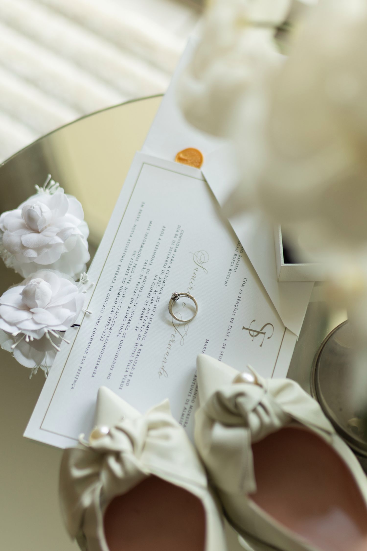 Elegant wedding flat lay with white shoes, ring, invitation card, and white roses on a mirrored surface.