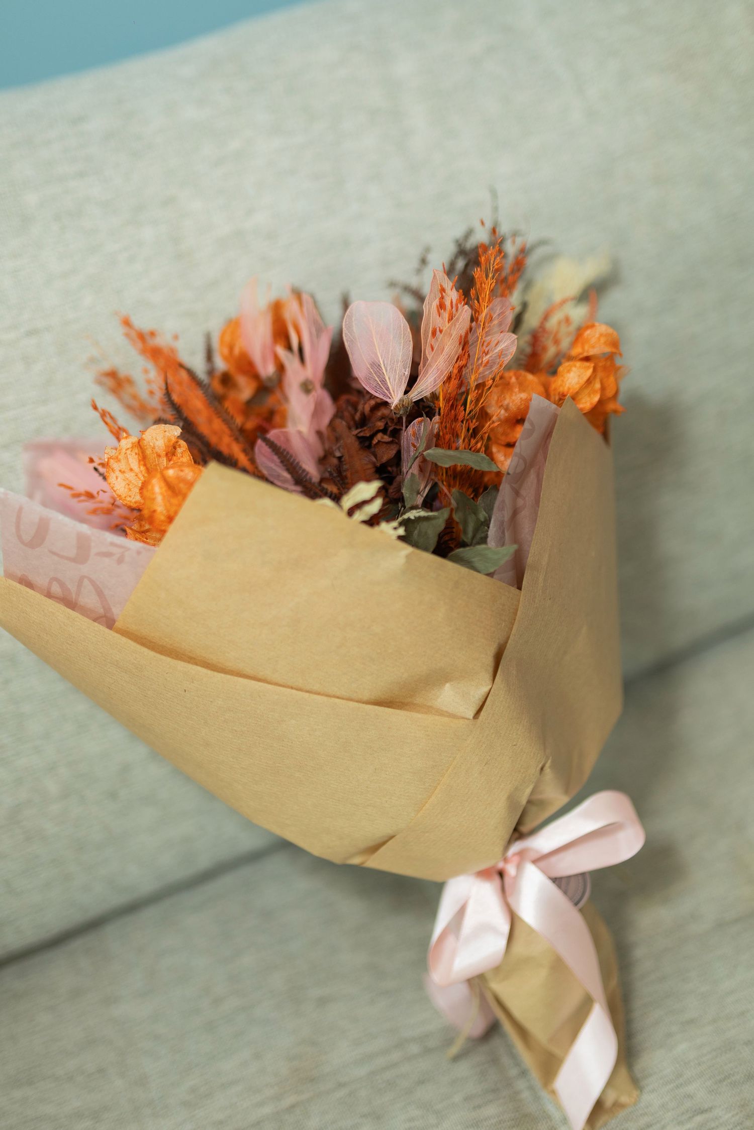 Dried flower bouquet wrapped in kraft paper with pink ribbon, featuring orange and pink blooms on a gray sofa.
