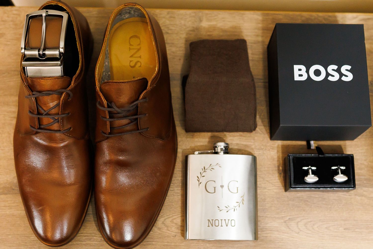 Brown leather dress shoes, Hugo Boss box, engraved flask, and cufflinks arranged as groom wedding accessories on wooden surface.