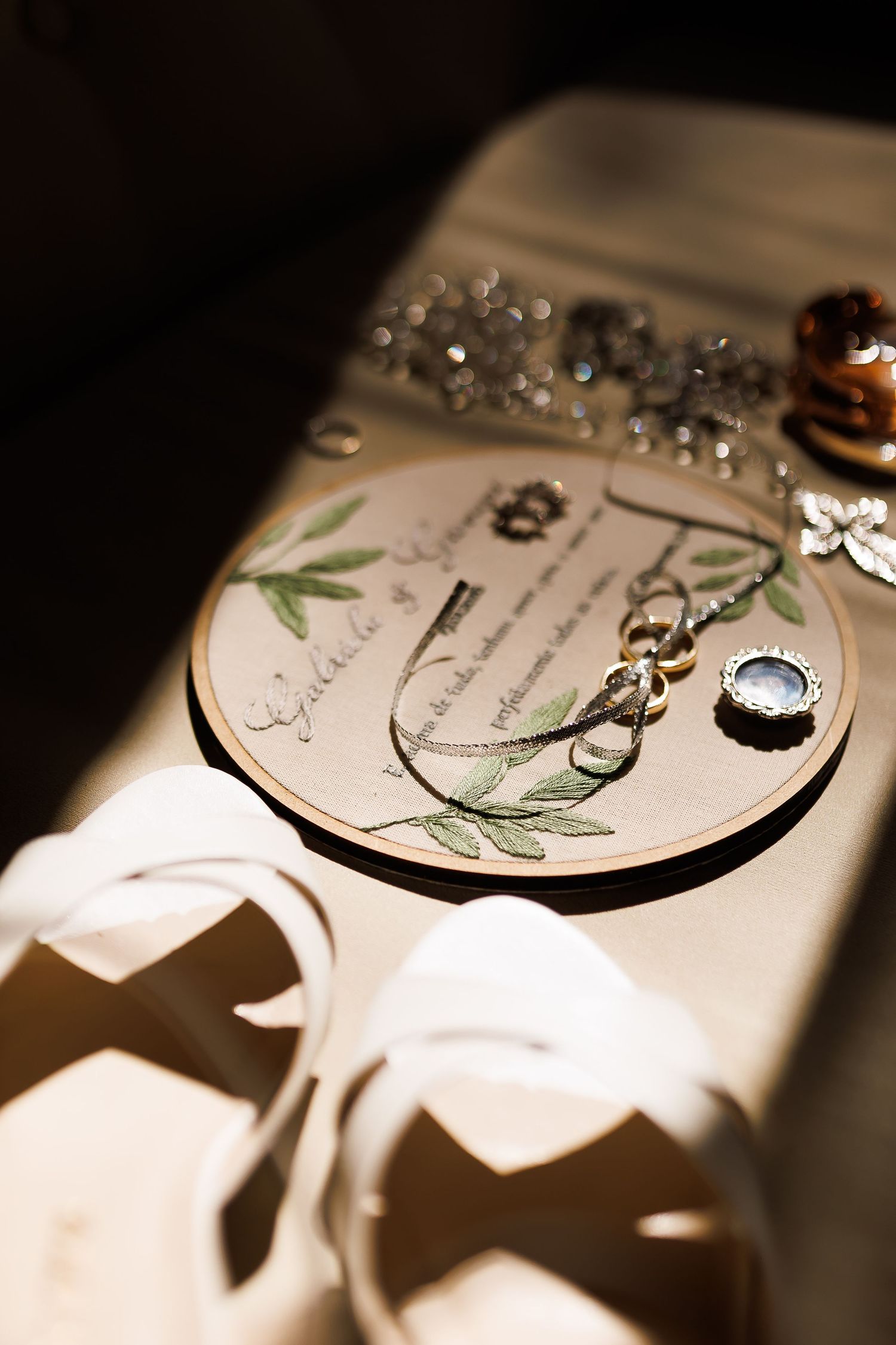 Elegant wedding ring display on embroidered hoop with botanical design, surrounded by jewelry and ribbons in soft natural light.