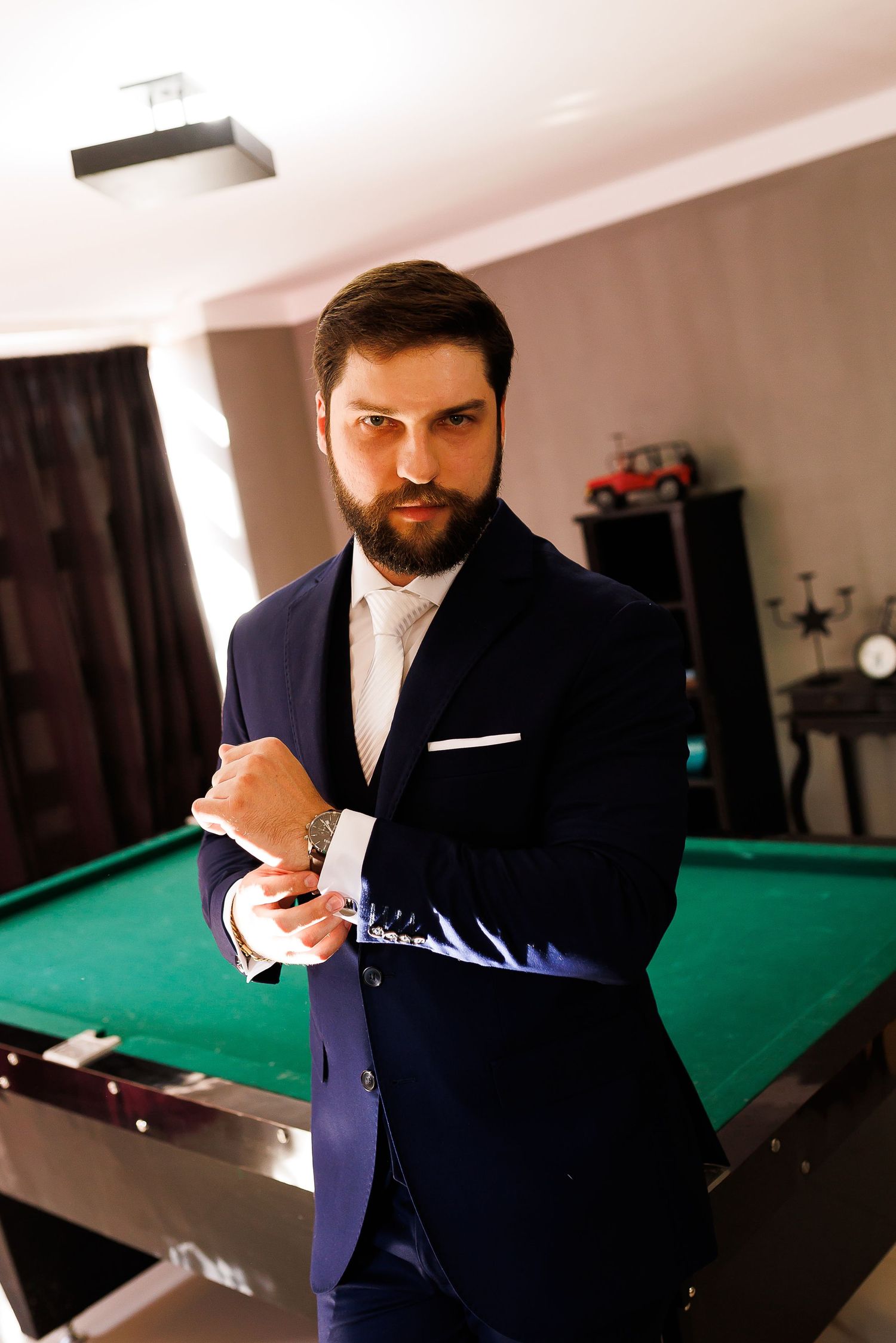 Bearded man in navy blue suit adjusting cufflinks beside a green billiard table in an elegant room.
