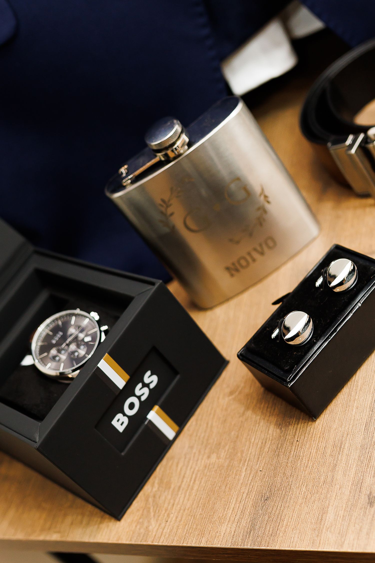 Hugo Boss watch in box, silver hip flask engraved NOIVO, cufflinks, and belt displayed on wooden surface.