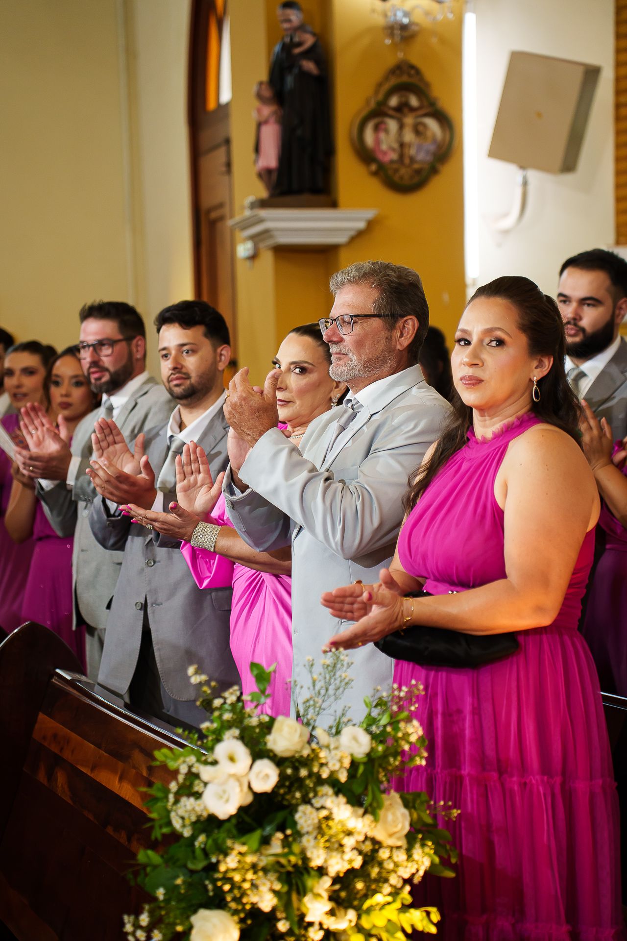 Wedding party in pink dresses and gray suits clapping in a church decorated with flowers and religious artwork.