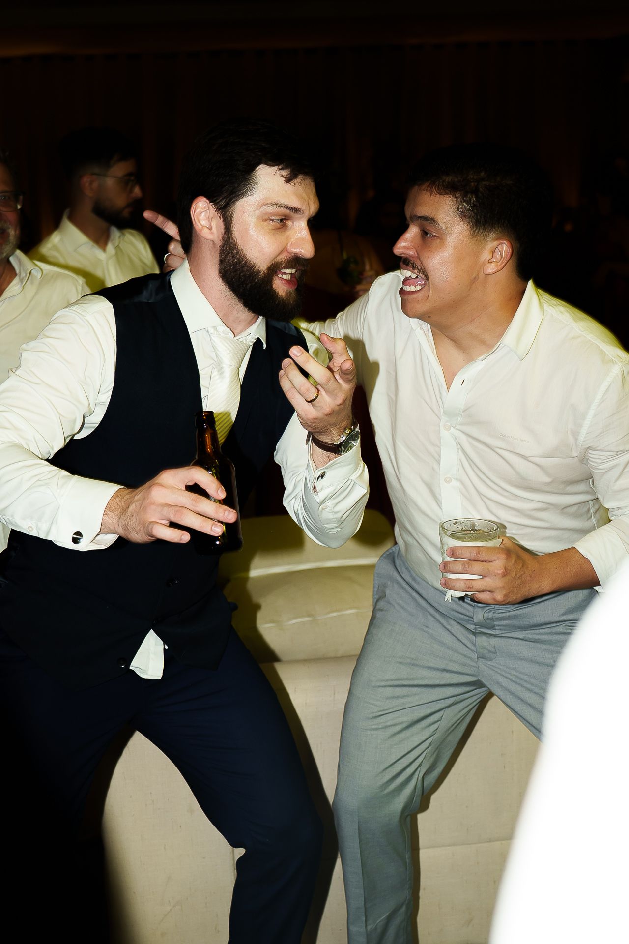 Two men in formal attire laugh and dance together at a party, each holding a drink.