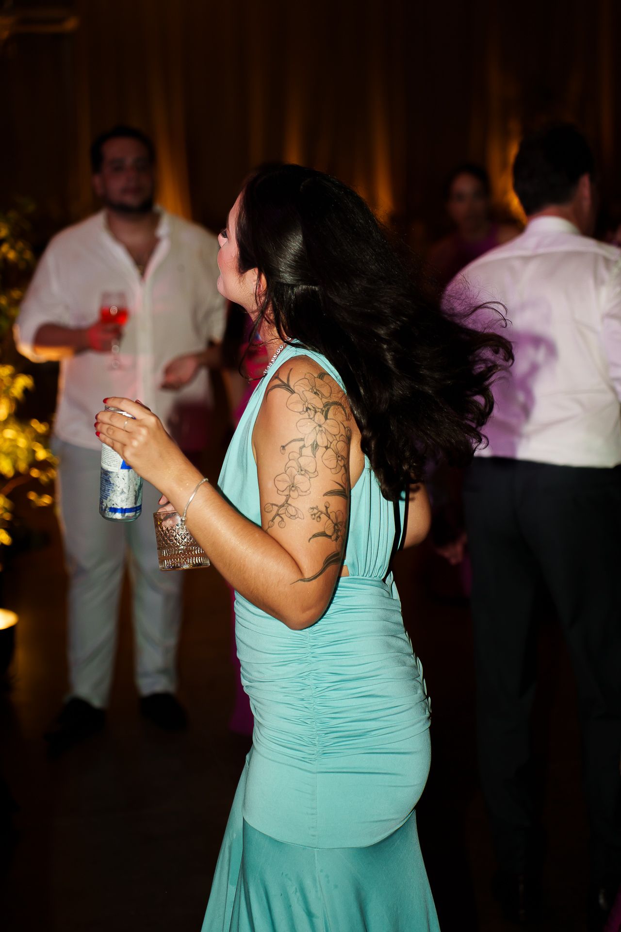 Woman in teal gown with back tattoos dancing at a wedding reception, holding a drink.