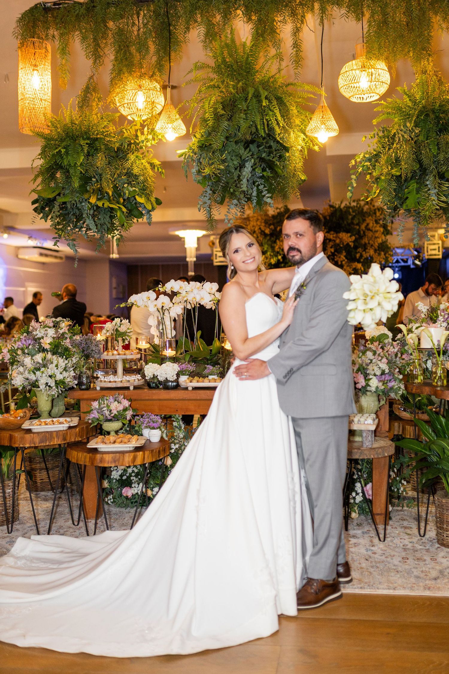 Bride in white gown and groom in gray suit embrace at elegant wedding reception with lush greenery and floral decor.